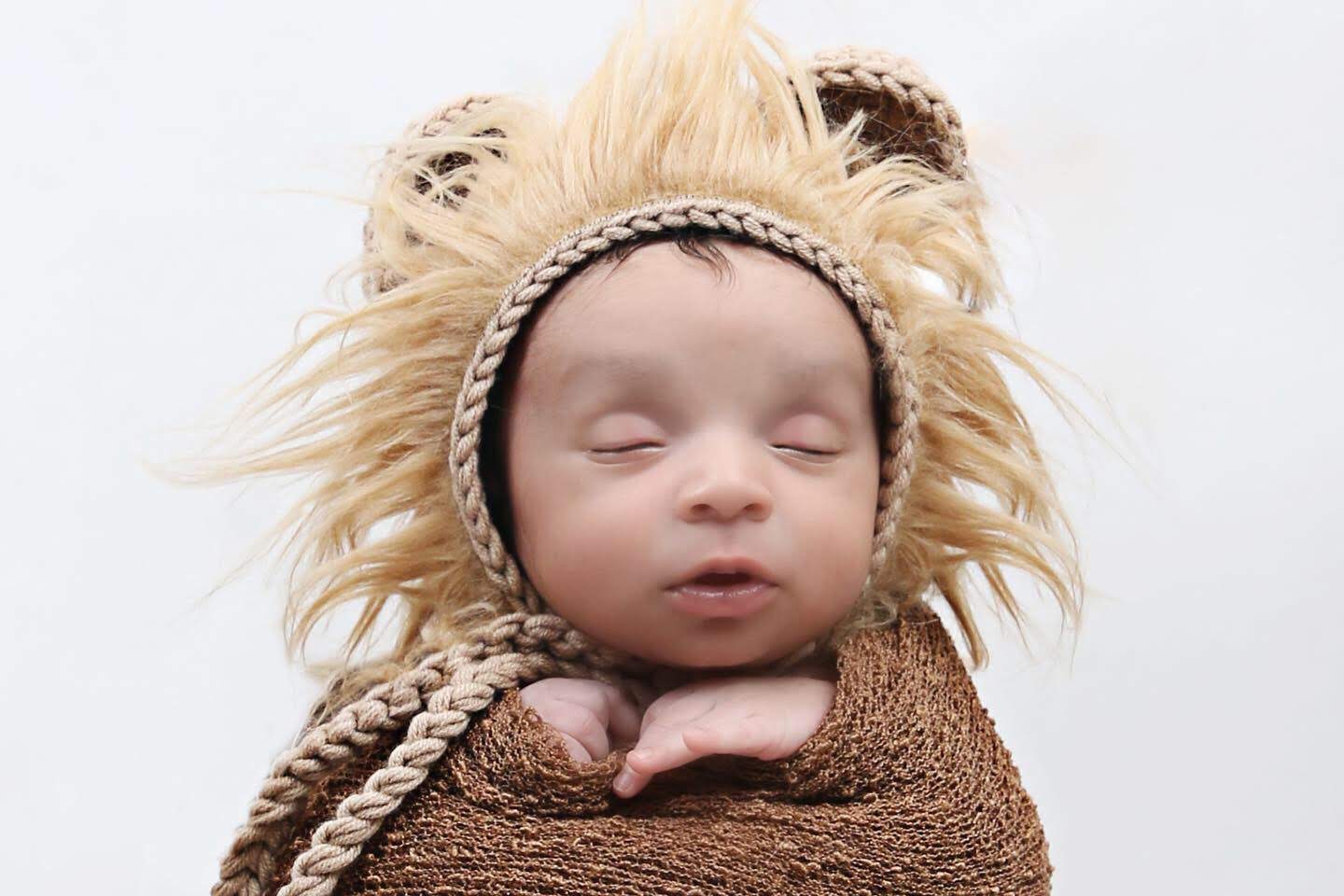 Sleeping newborn in a fluffy knit lion bonnet and brown wrap