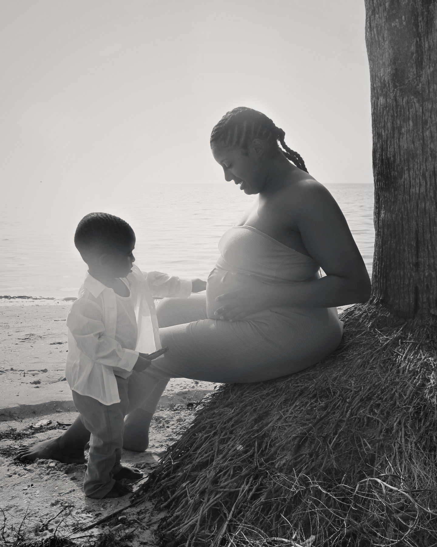 Black-and-white maternity photograph of a pregnant mother seated by a tree at the beach with her young son