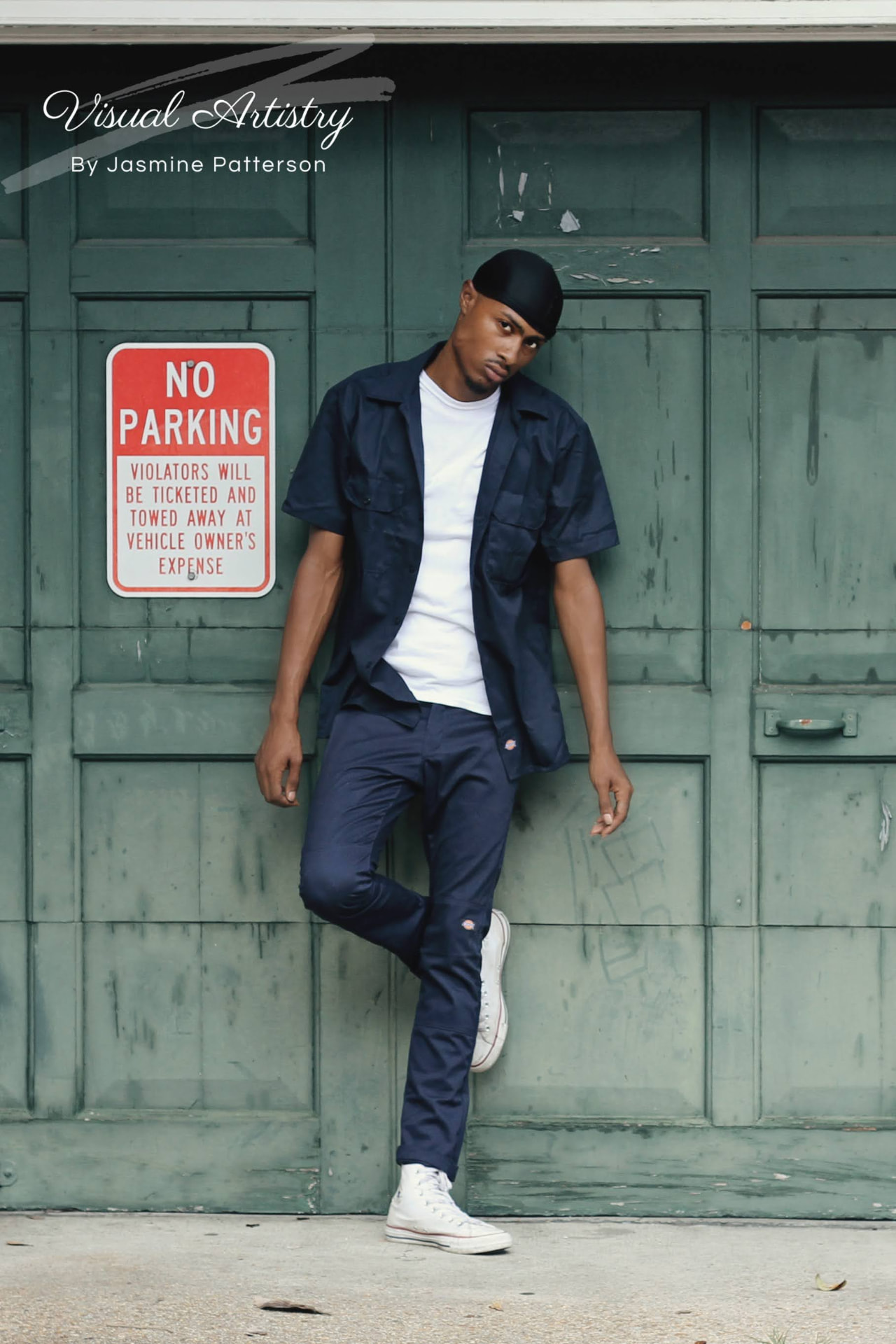Young man in navy leaning against weathered green doors beside a no parking sign