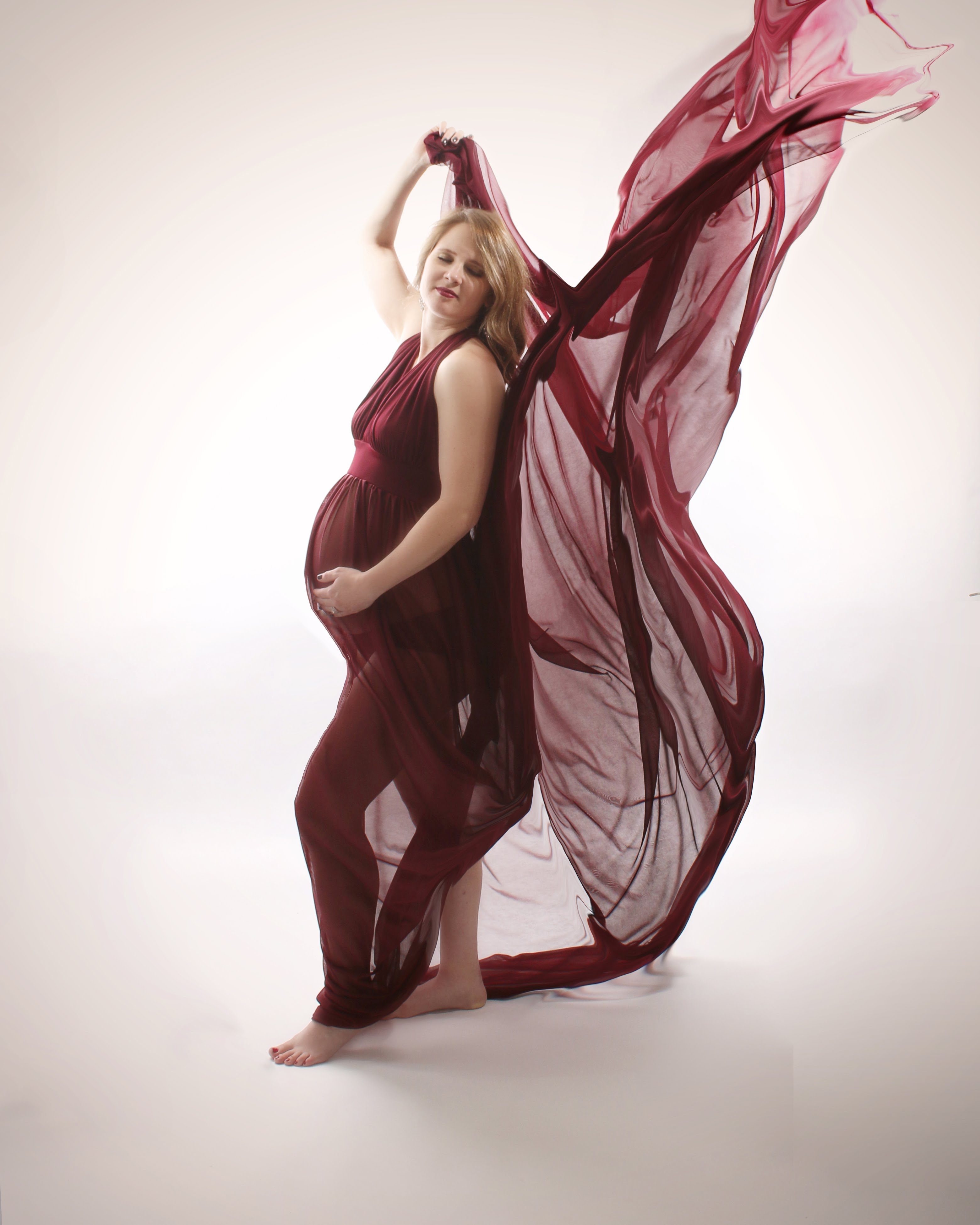 Expectant mother in a burgundy sheer gown with arms raised on a white studio backdrop