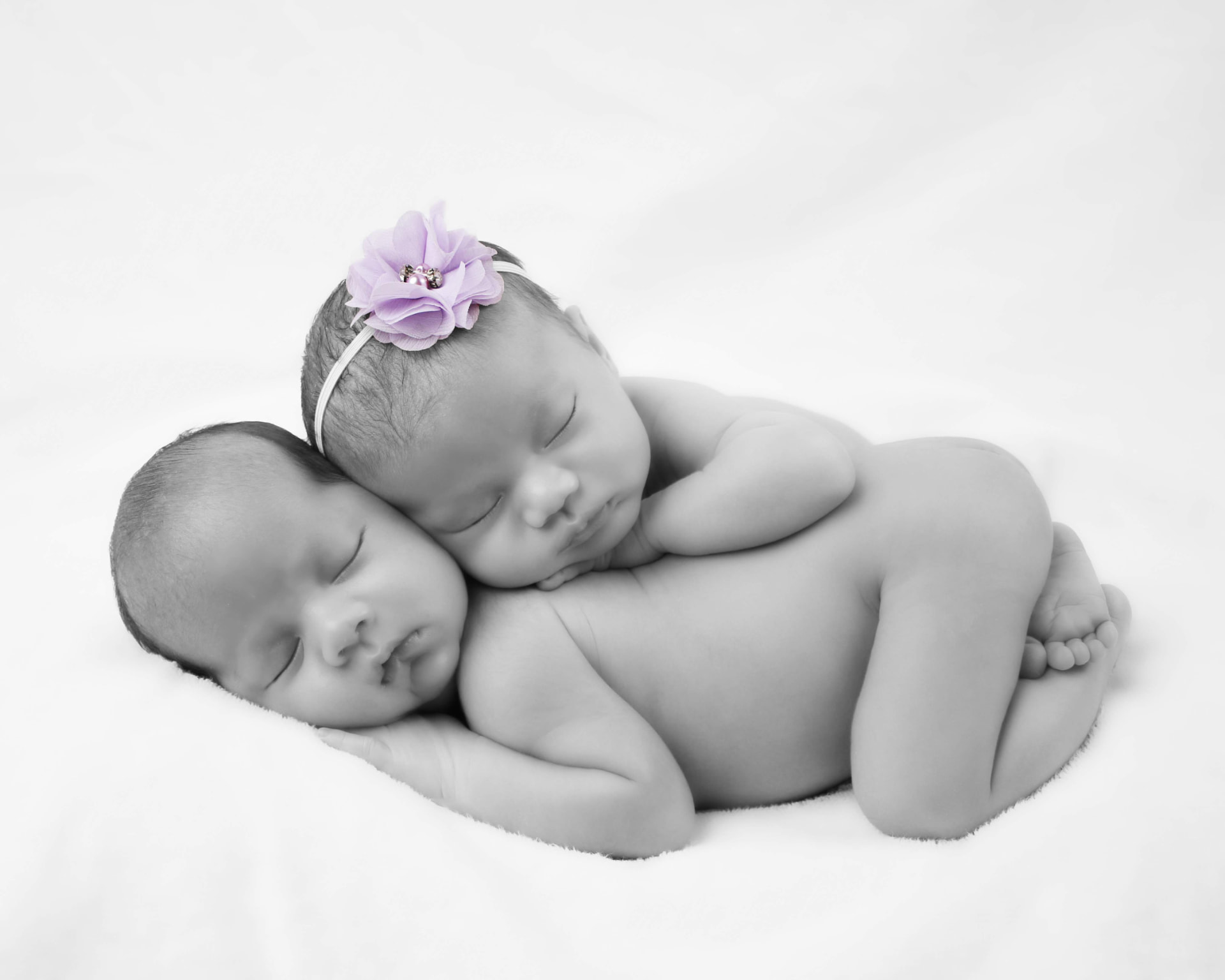 Newborn twins sleeping together, one with a lavender flower headband, black and white