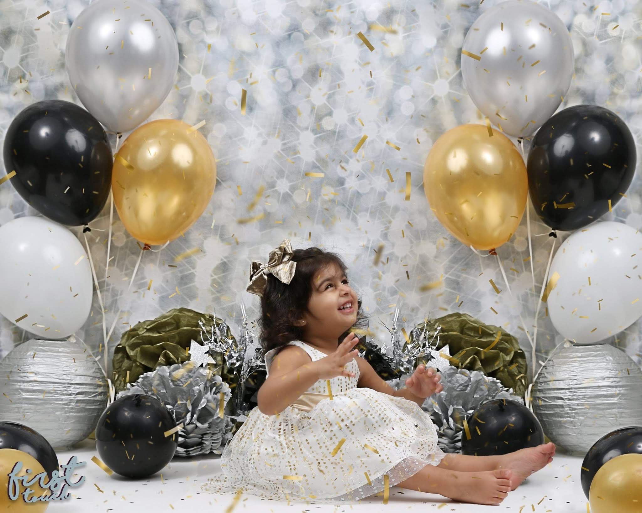 Toddler in a white dress seated among black, white, and gold birthday balloons