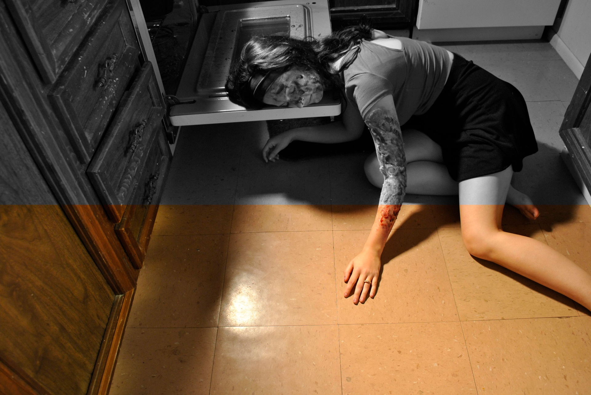 Woman lying on a kitchen floor beside an open oven, rendered mostly in black and white with selective warm color