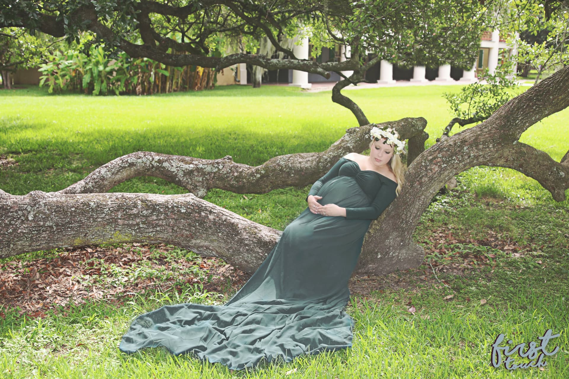Pregnant woman in a long green gown and flower crown leaning on a sprawling oak limb