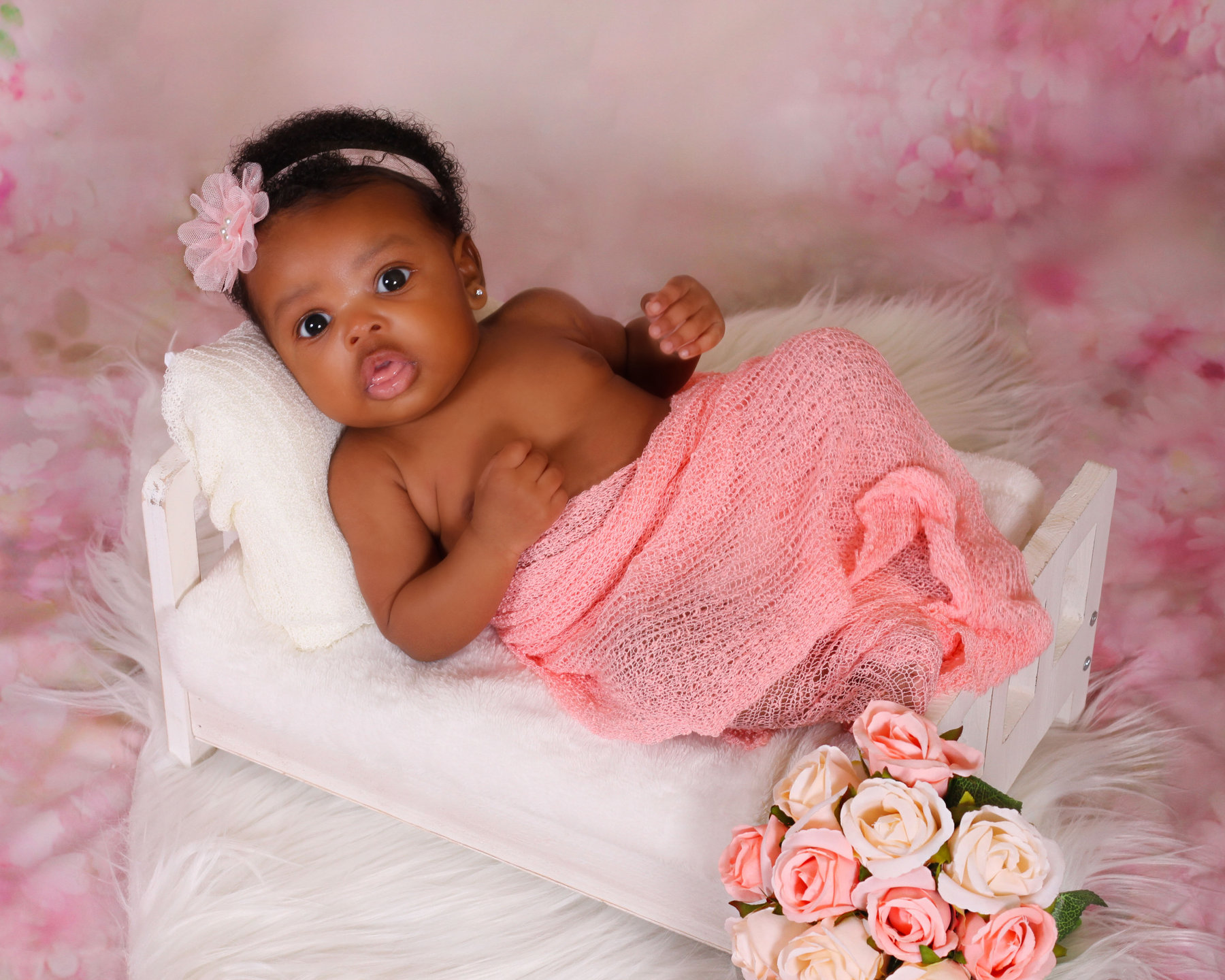 Newborn girl with a flower headband lying on a pink backdrop with roses