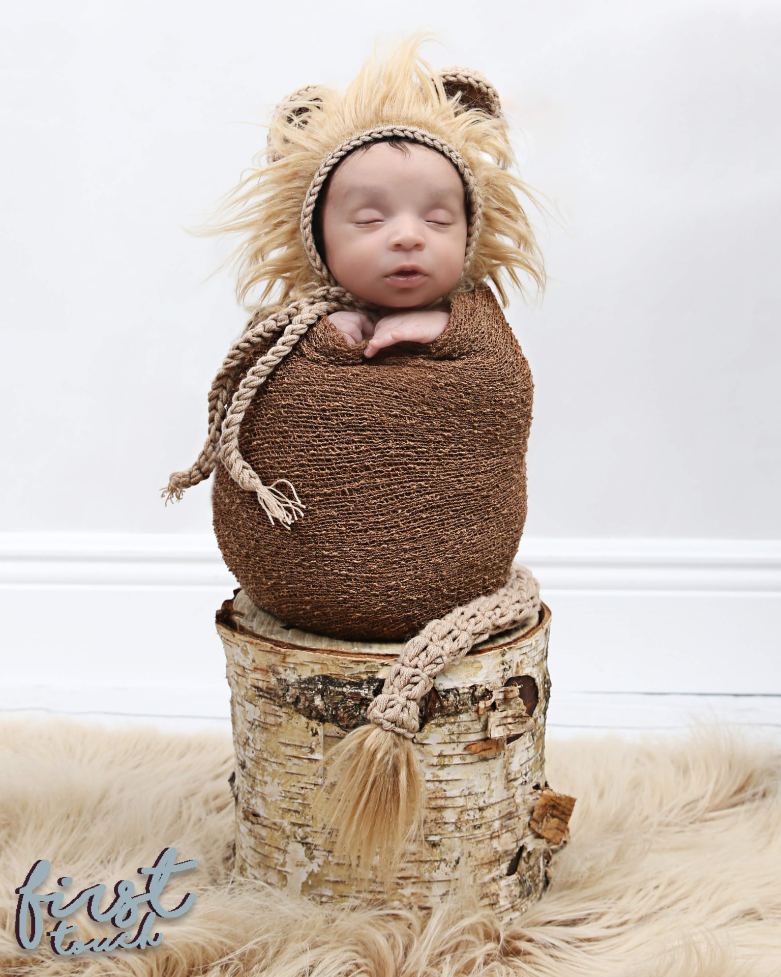 Newborn baby in a knit lion hat resting on a birch-log pedestal over a fur rug