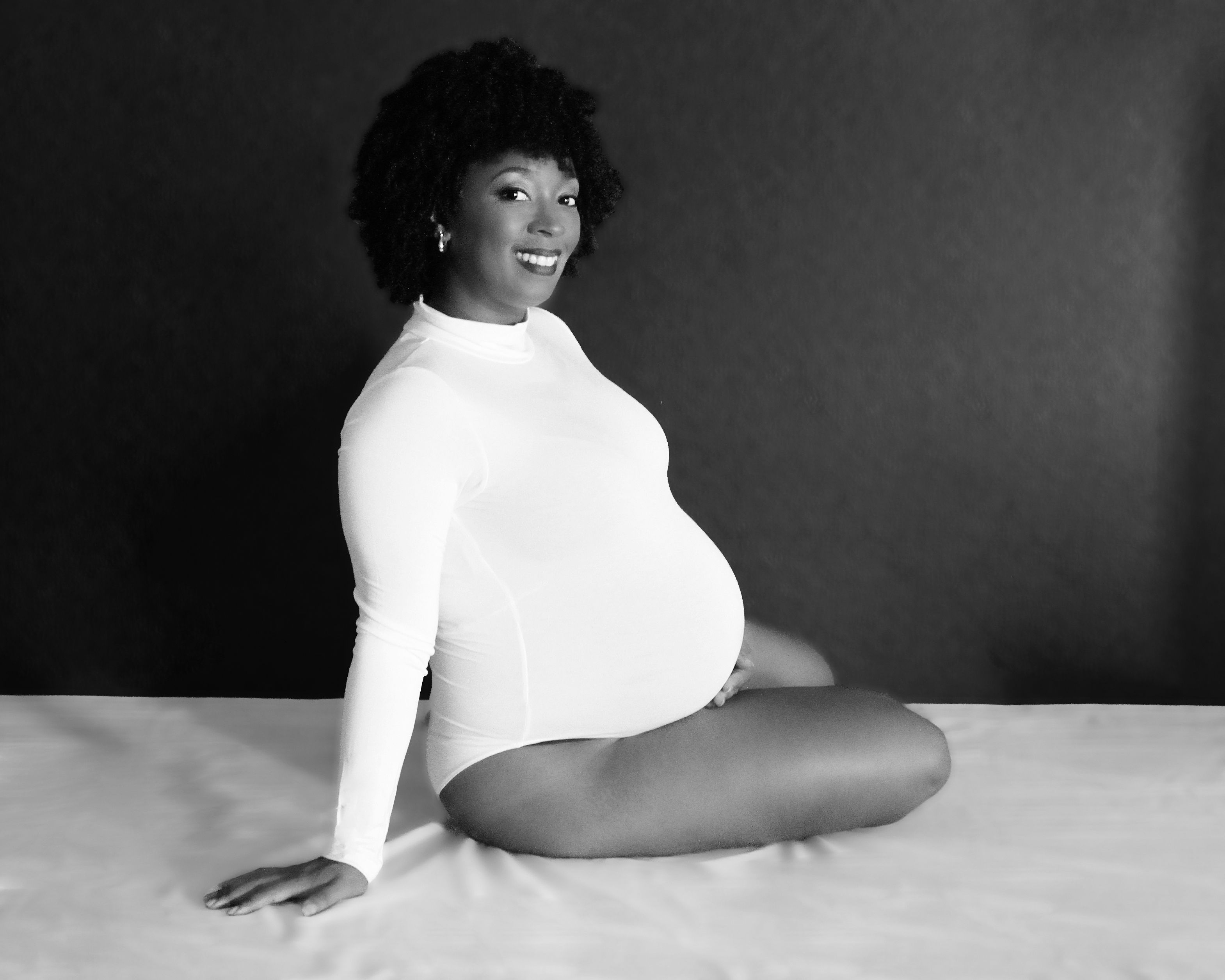 Black-and-white studio maternity portrait of an expectant mother seated in a white bodysuit