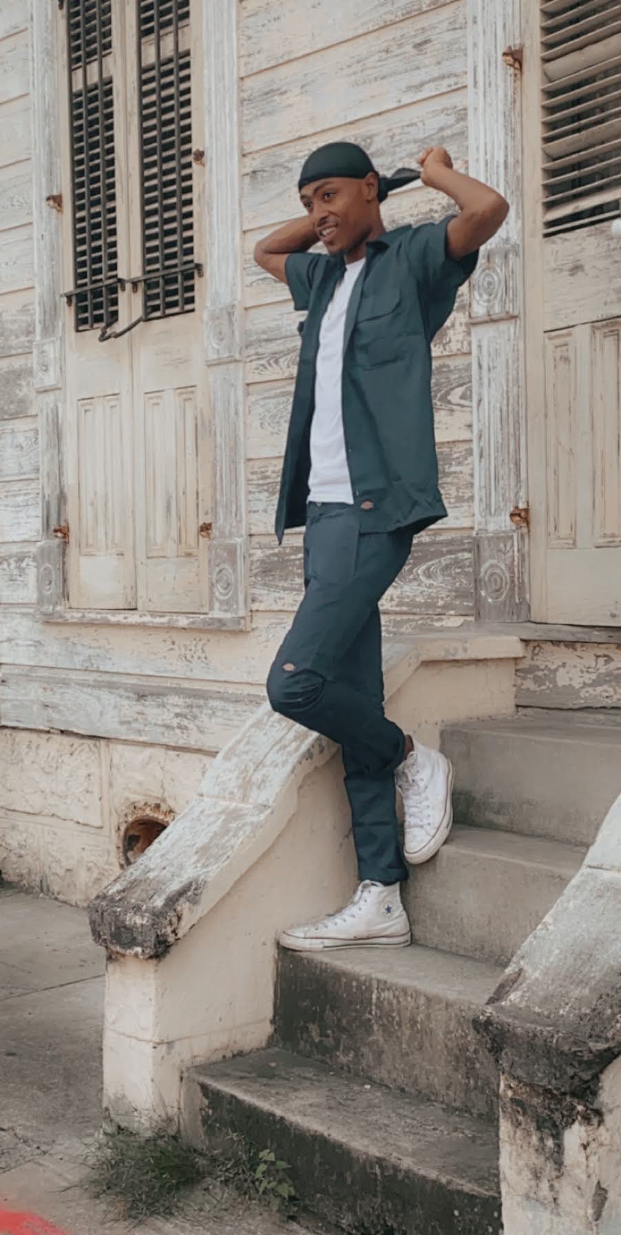 Young man in a dark green outfit and white sneakers standing on the steps of a weathered cottage