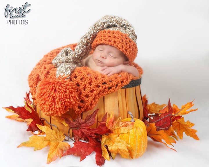 Newborn in an orange knit bonnet curled in a basket with fall leaves and pumpkin ornaments