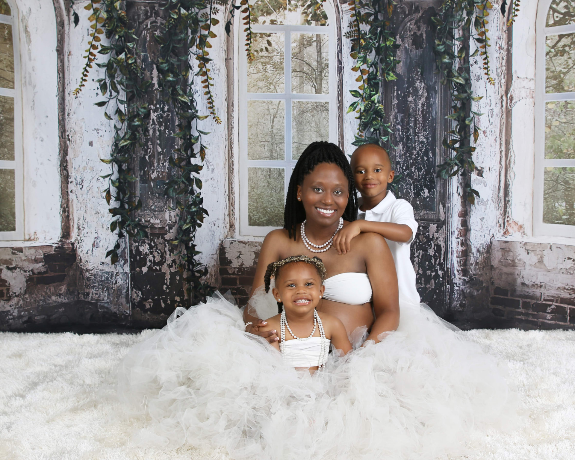 Pregnant mother in white seated on a fur rug with her two children in a rustic window-lit studio set