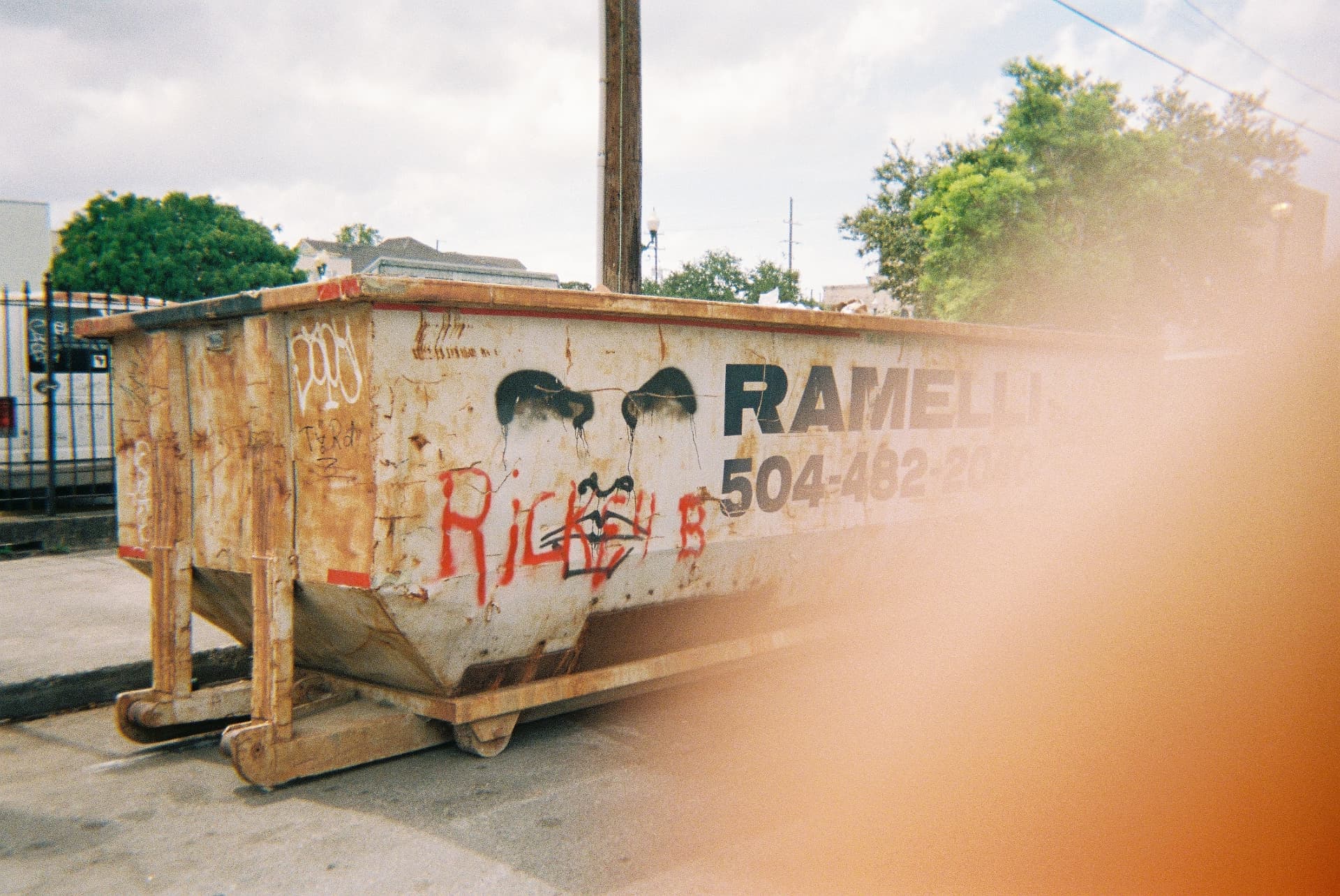 Weathered, graffiti-marked dumpster with faded eyes painted on it, under an orange film light leak