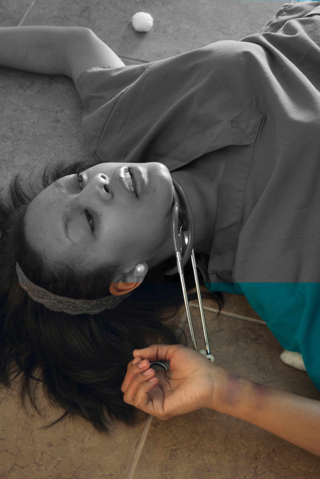 Woman lying on a tiled floor with a stethoscope around her neck, rendered mostly in black and white with selective teal color