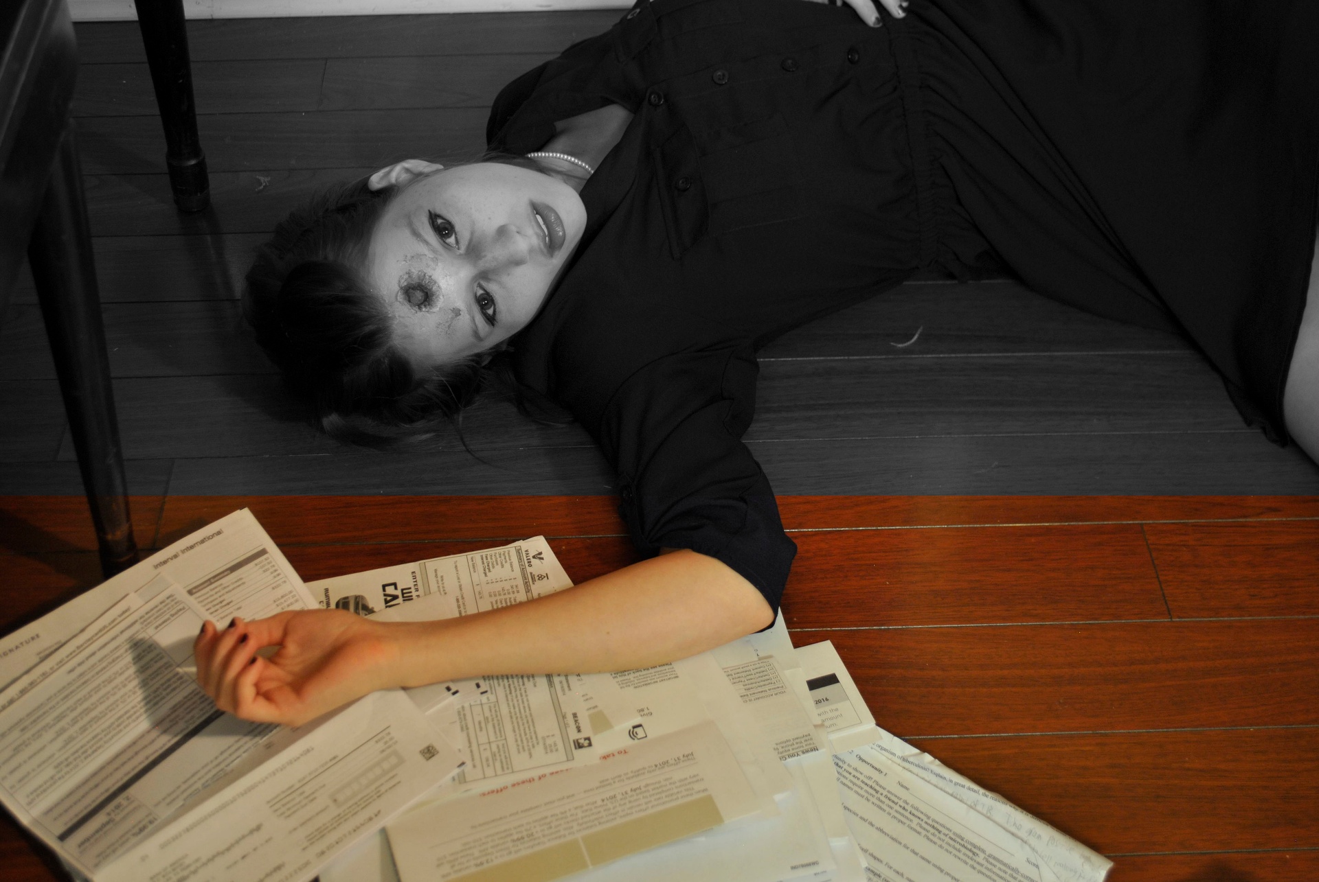 Businesswoman lying among scattered office papers in a staged gunshot scene, rendered mostly in black and white with selective warm color