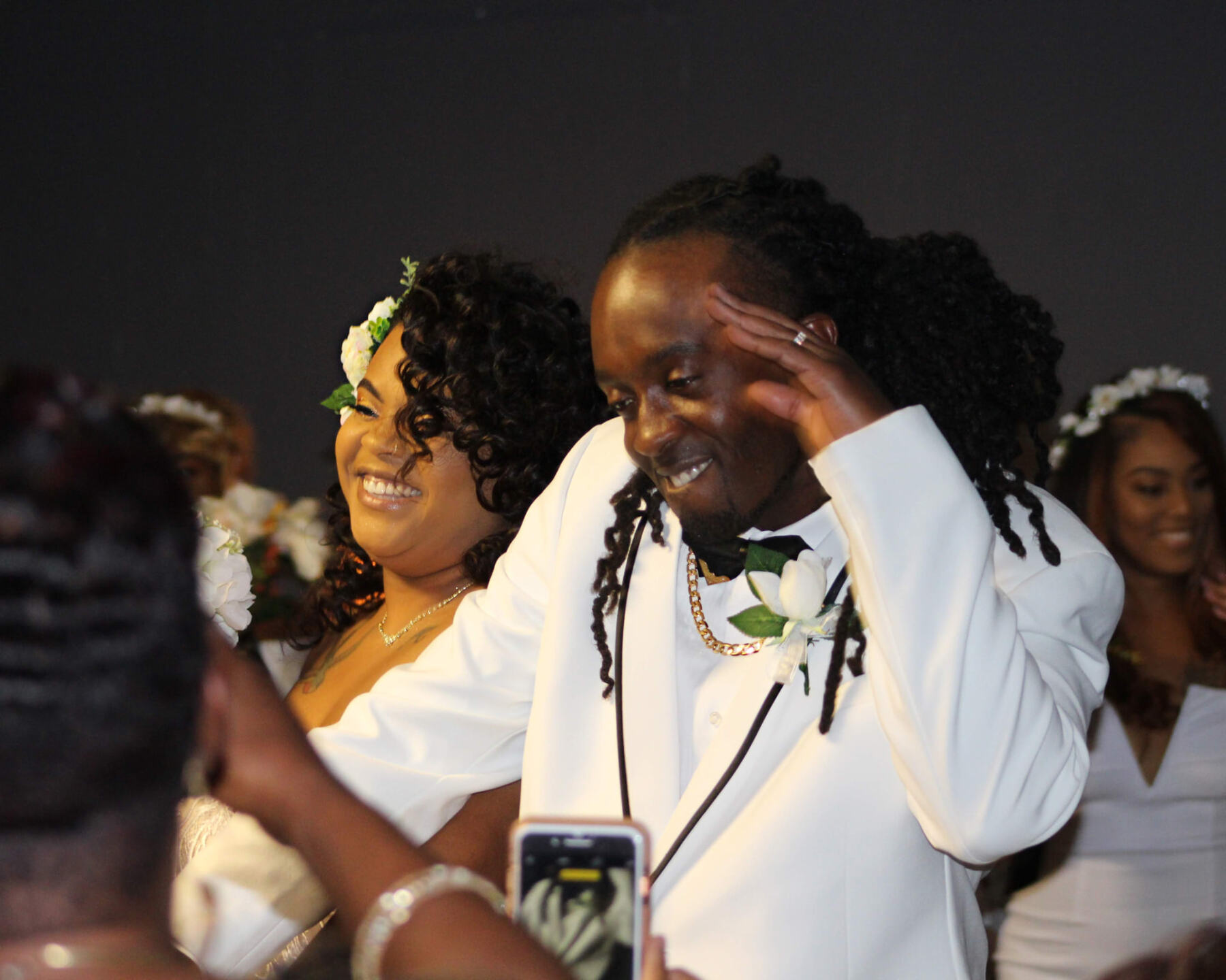 Groom in a white tuxedo grinning and saluting as the bride laughs beside him