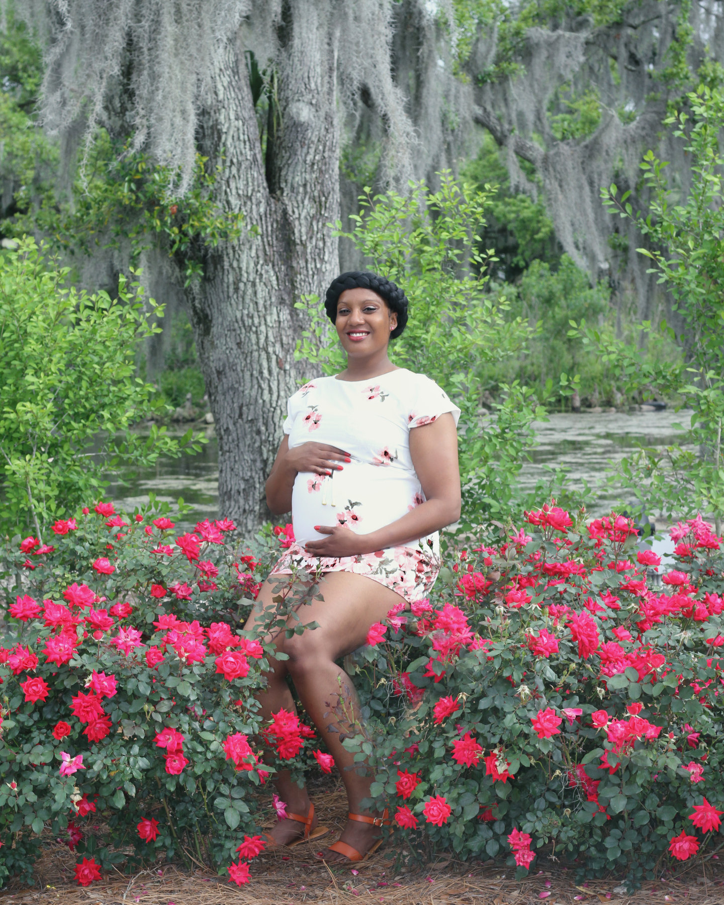 Pregnant woman seated among red flowers beneath a moss-draped oak tree