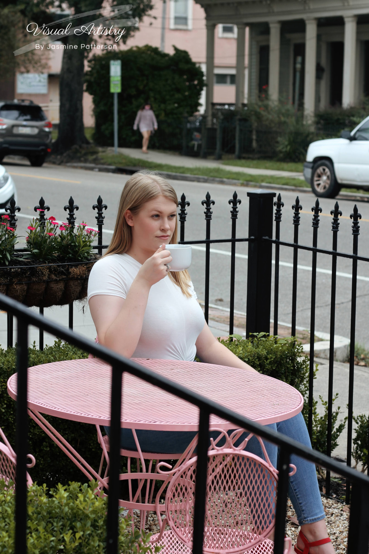 Woman in a white top seated at a pink café table on the sidewalk, holding a cup