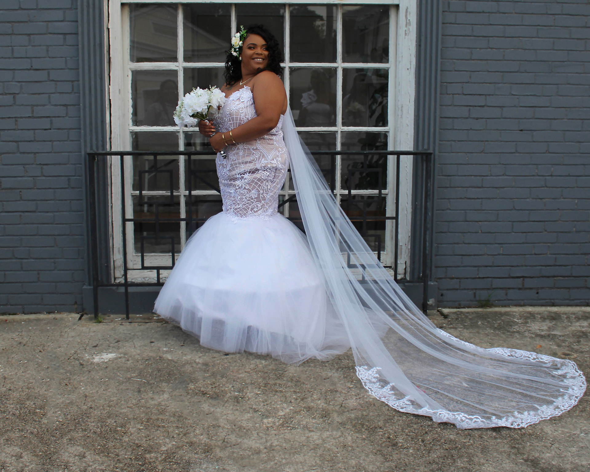 Bride in a lace mermaid gown with a long veil and flower crown outside a grey brick building