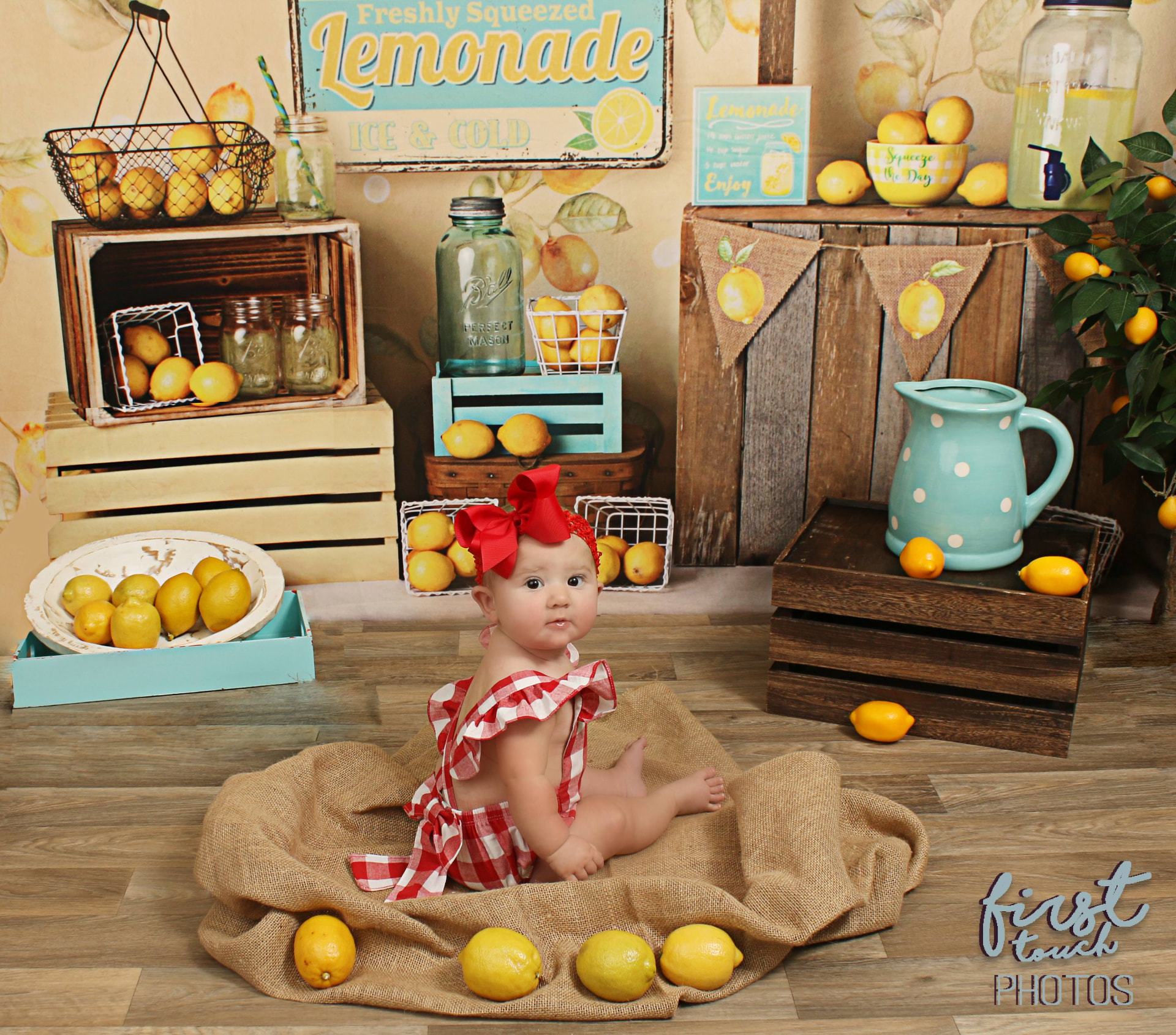 Baby in a red gingham romper and bow sitting on burlap in a lemonade stand set