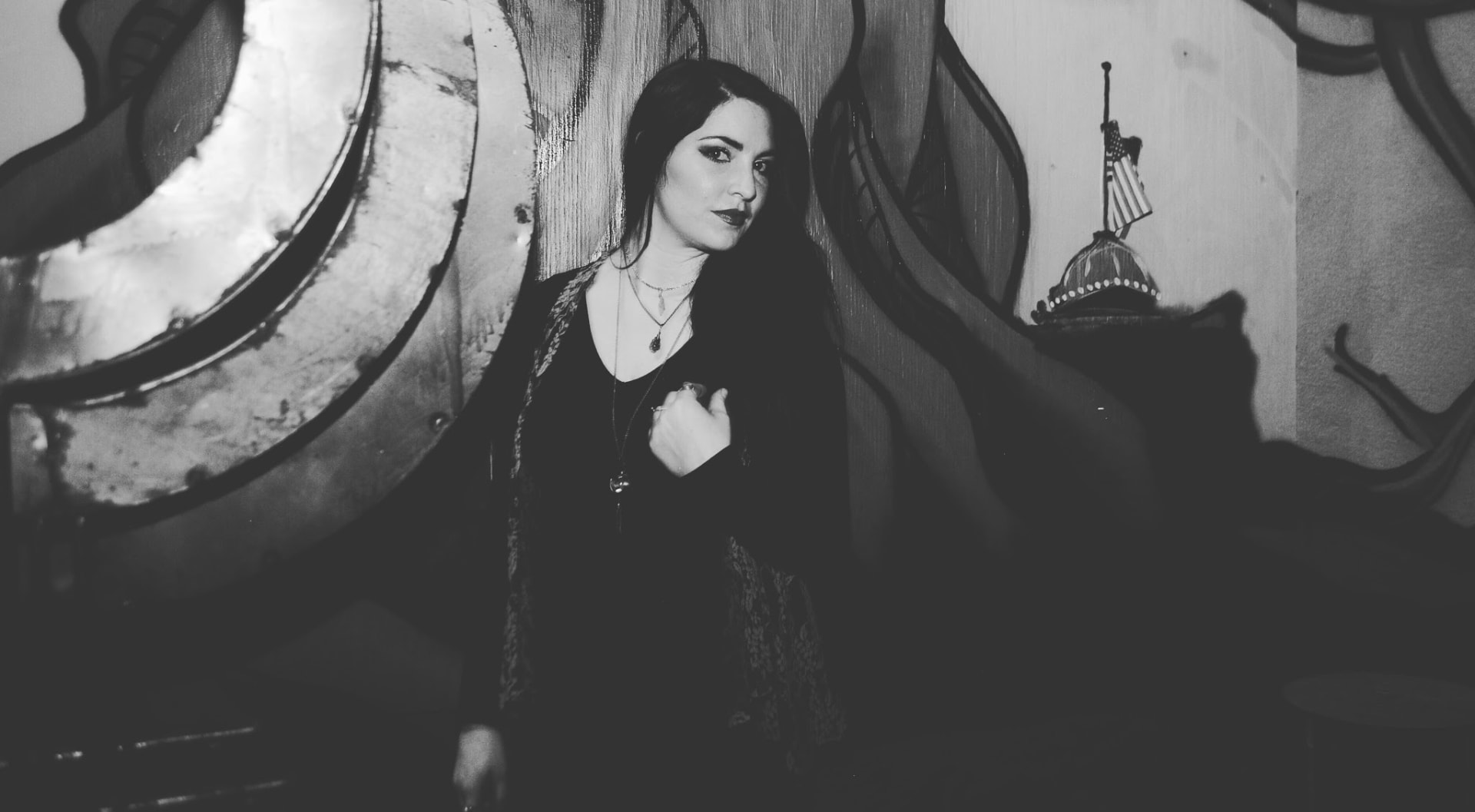 Black-and-white portrait of a dark-haired woman in black standing before a mural