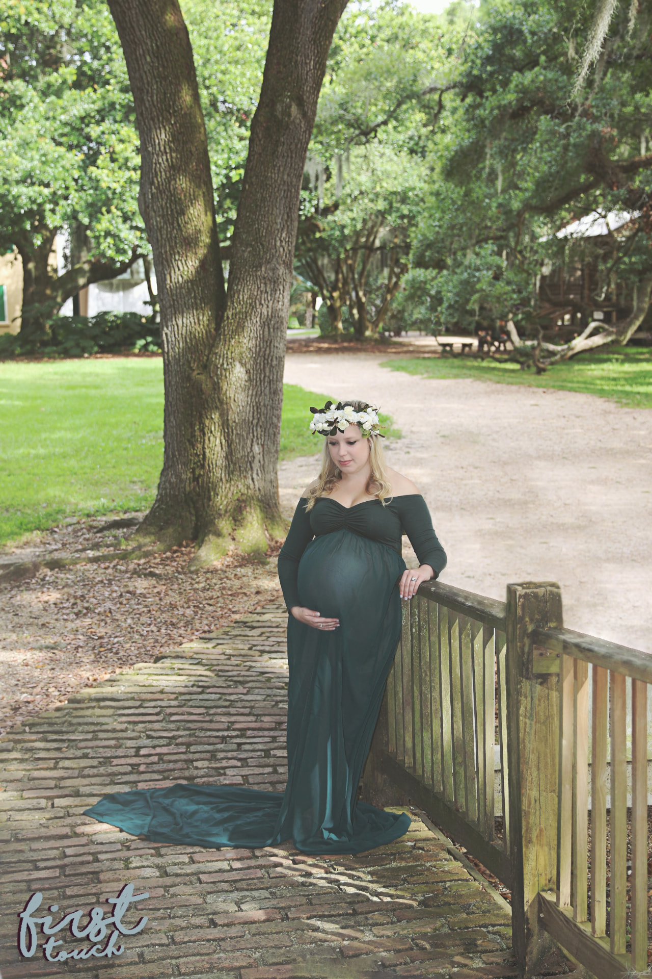 Maternity portrait in a long emerald gown and white flower crown beside a wooden fence under live oaks