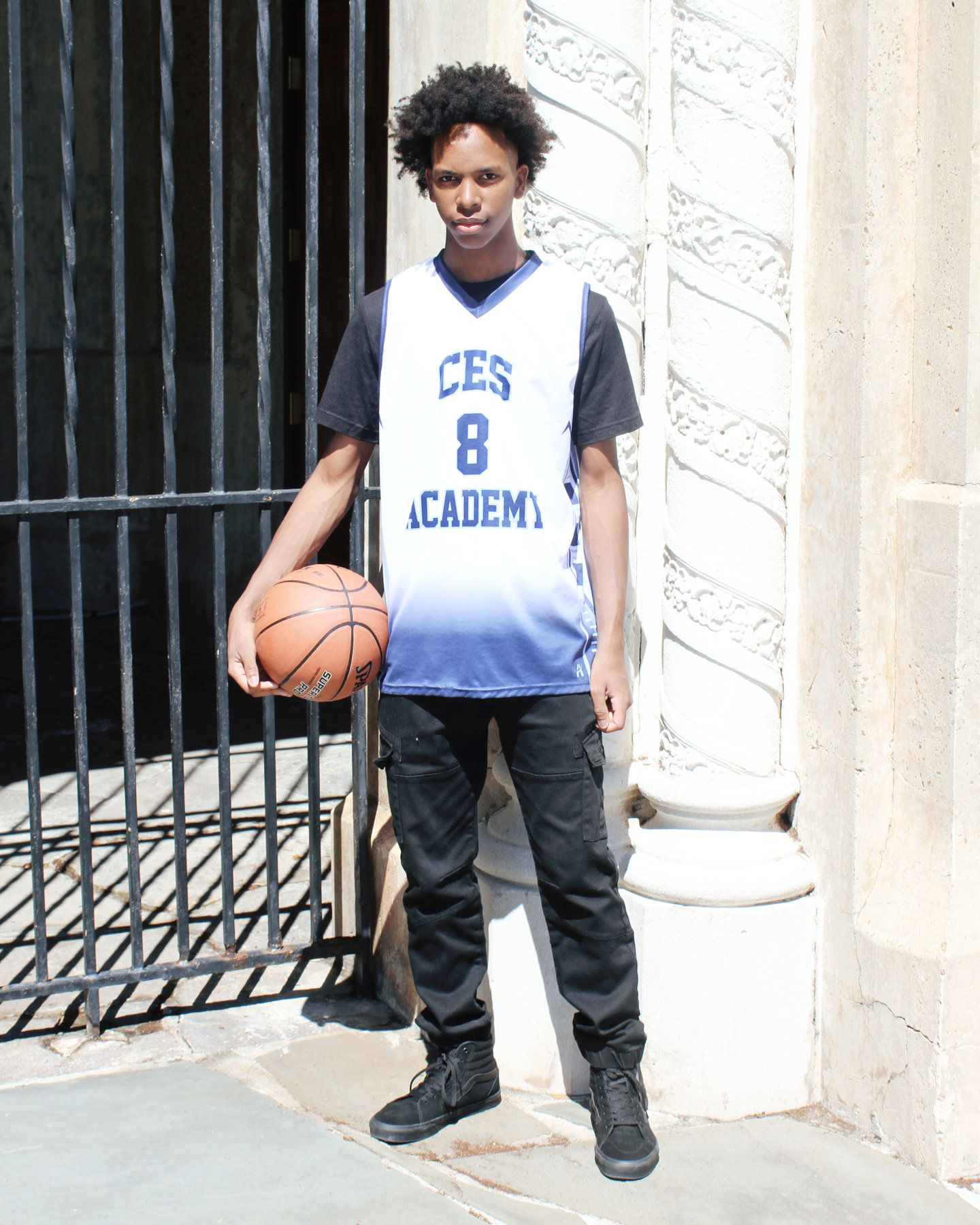 Teen in a basketball jersey holding a basketball beside an ornate gate