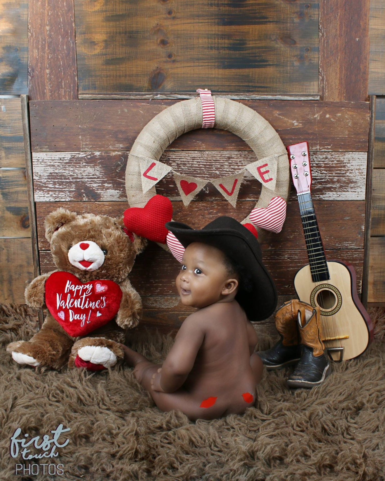 Baby in a black cowboy hat on a fur rug beside a teddy bear, guitar, and LOVE wreath
