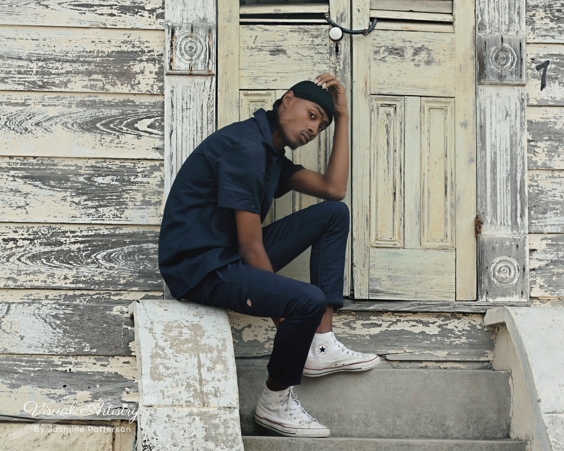 Young man in a navy outfit seated on the steps of a weathered Creole cottage, hand on his cap