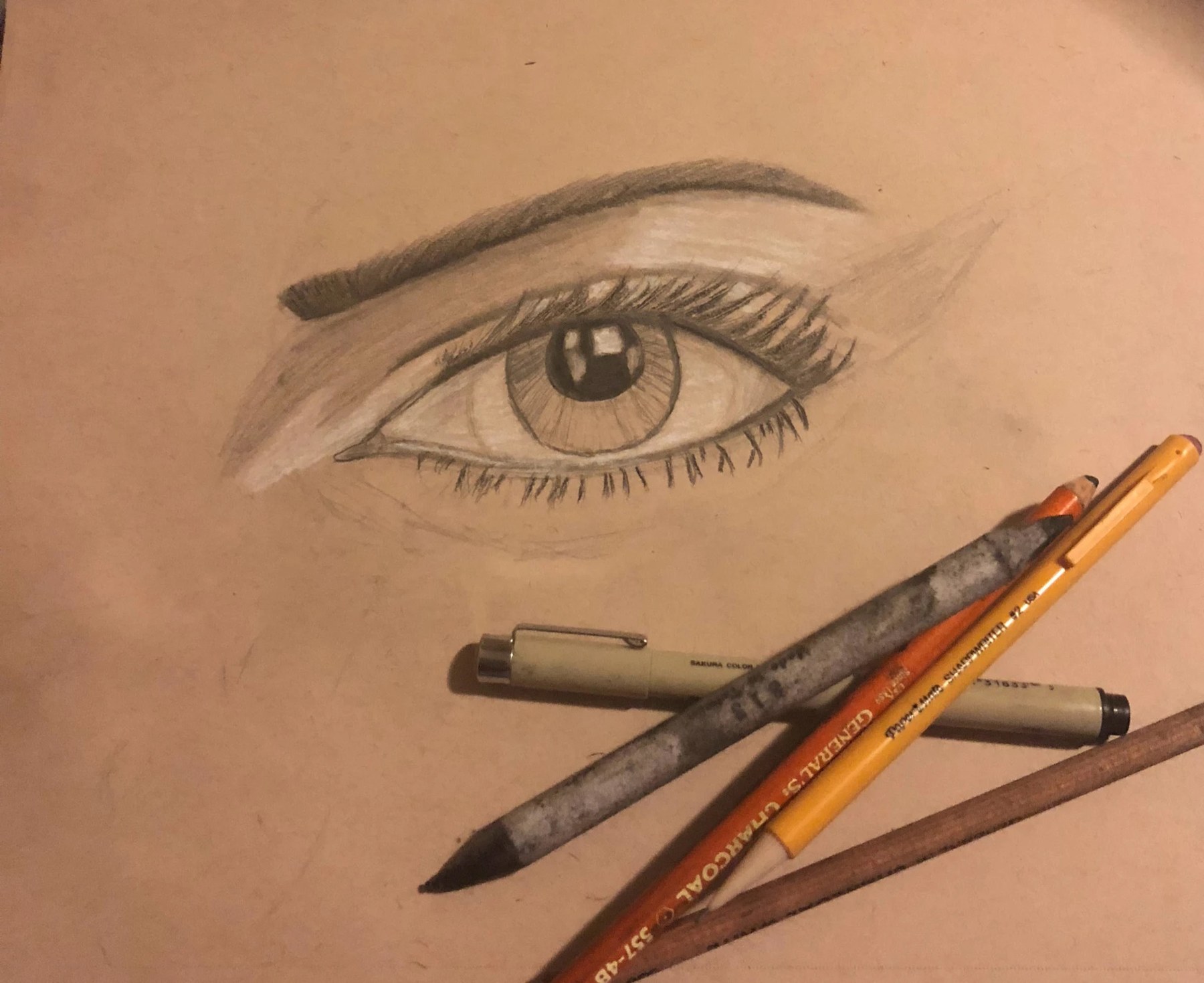Graphite drawing of a realistic eye on toned paper with drawing pencils and a pen laid beside it