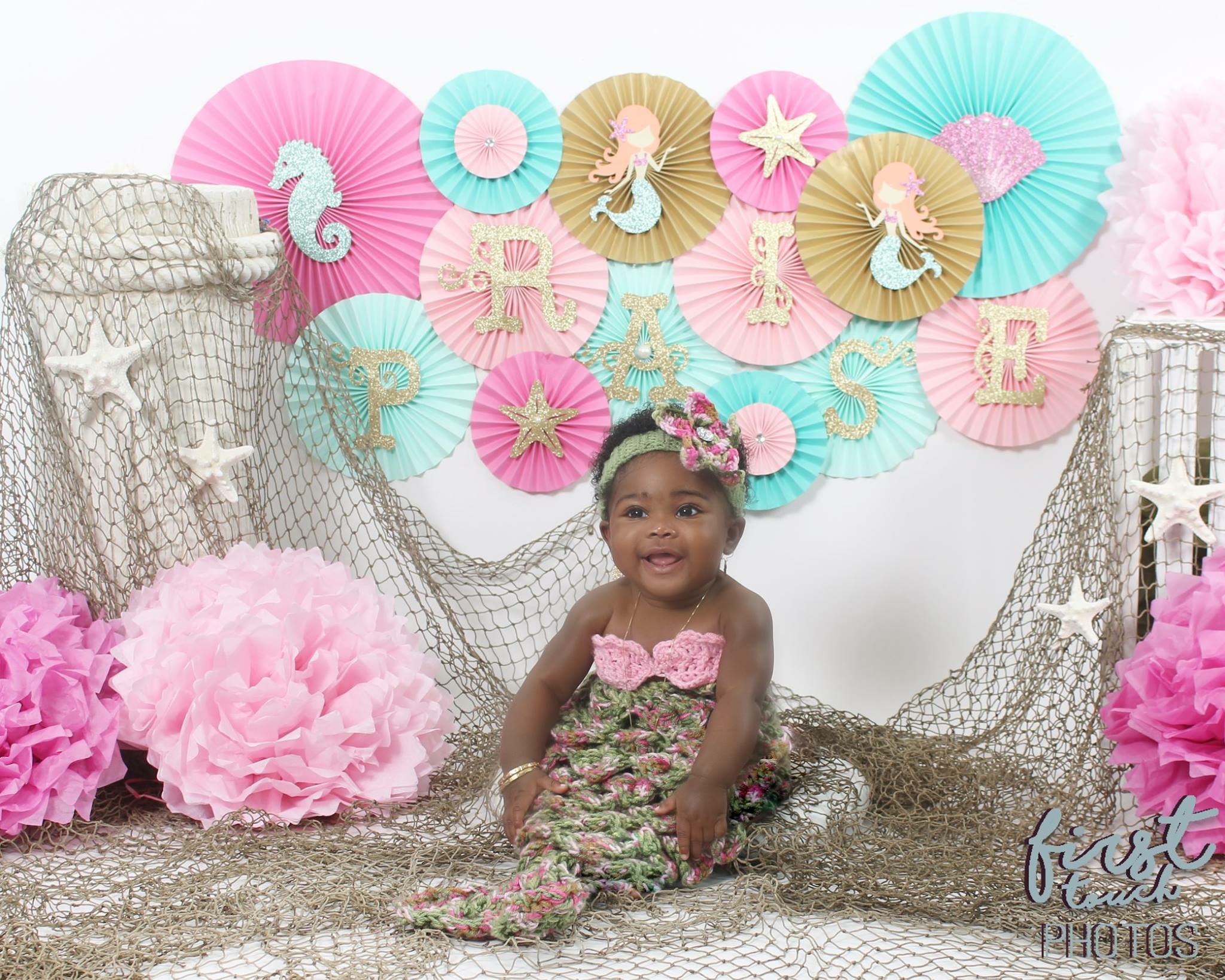 Baby in a mermaid tail set beneath a fan of mermaid paper rosettes and fishing net