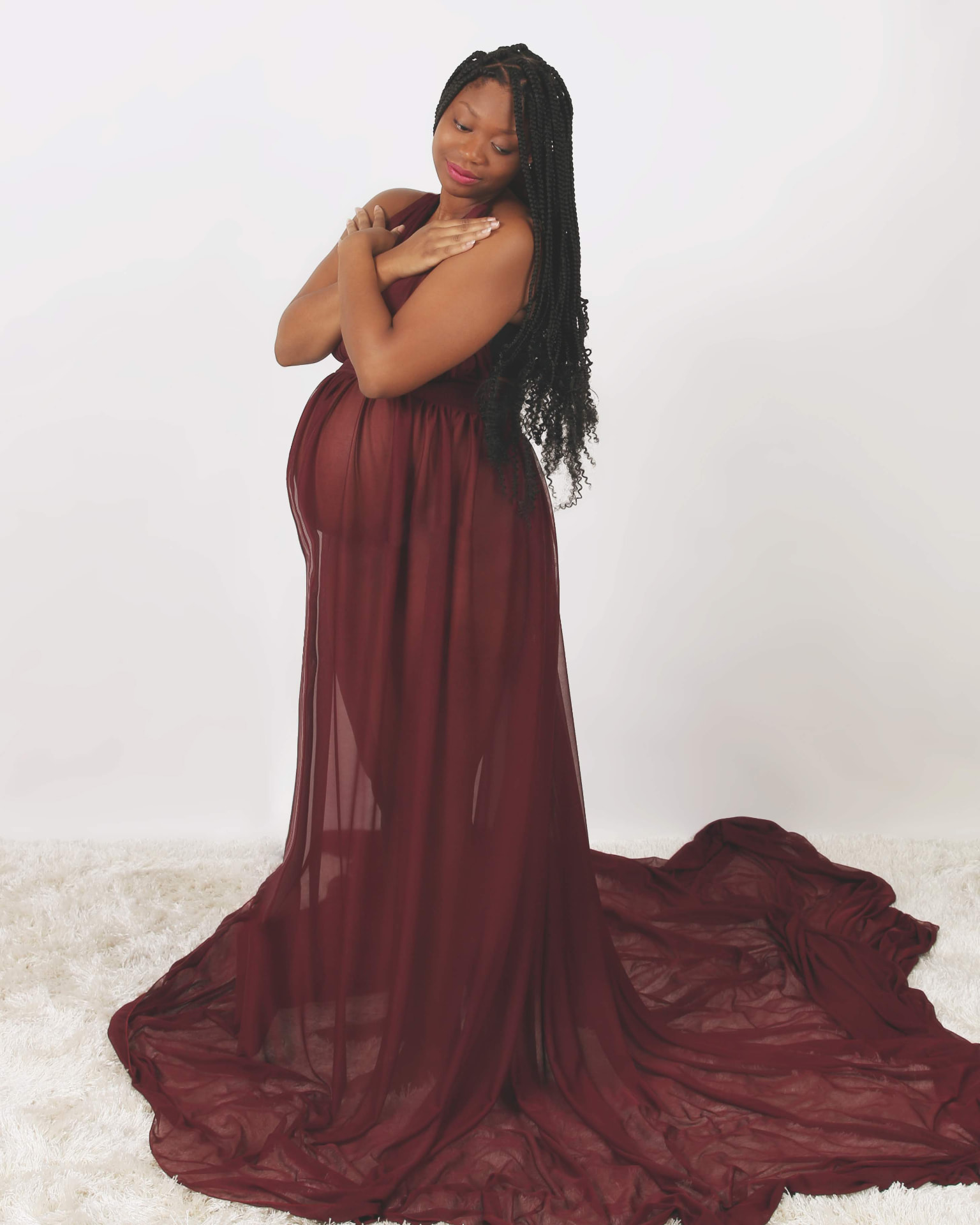Pregnant woman in a maroon gown with arms crossed, standing on a white fur rug in a bright studio