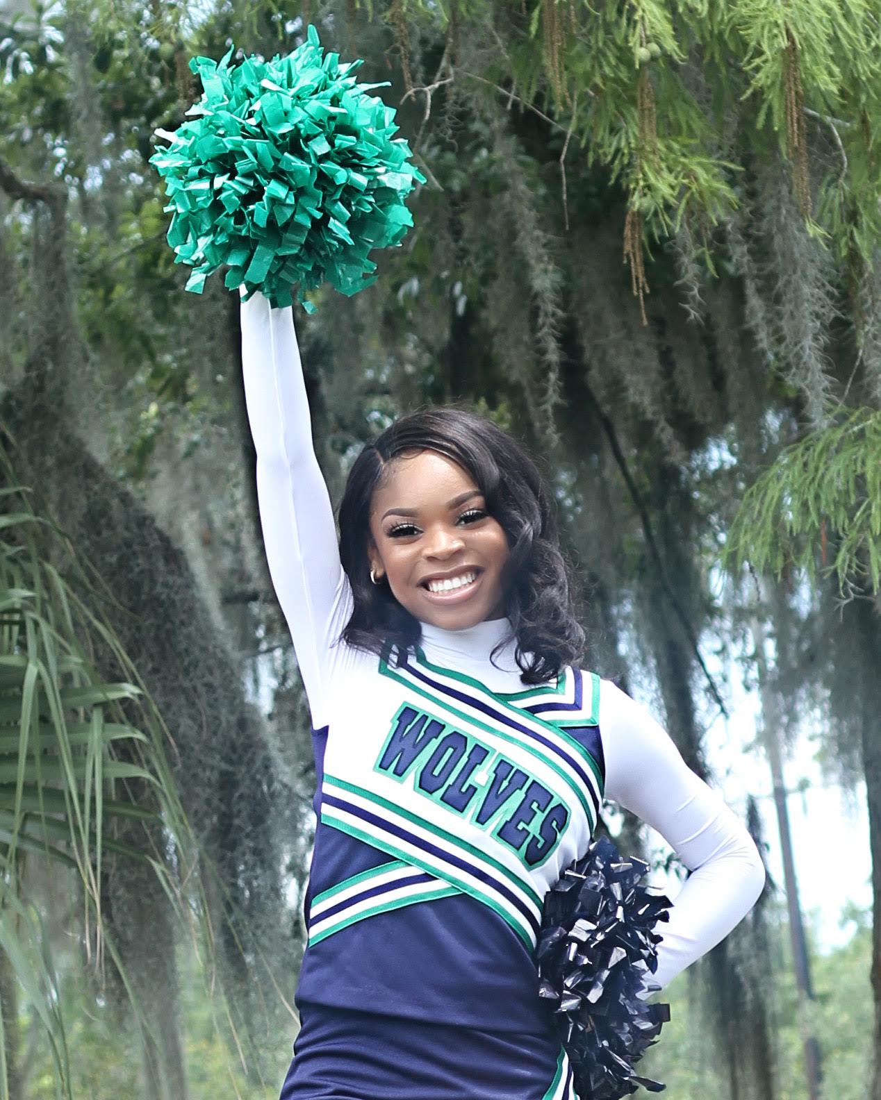 Cheerleader in a WOLVES uniform raising a green pom-pom against moss-draped trees