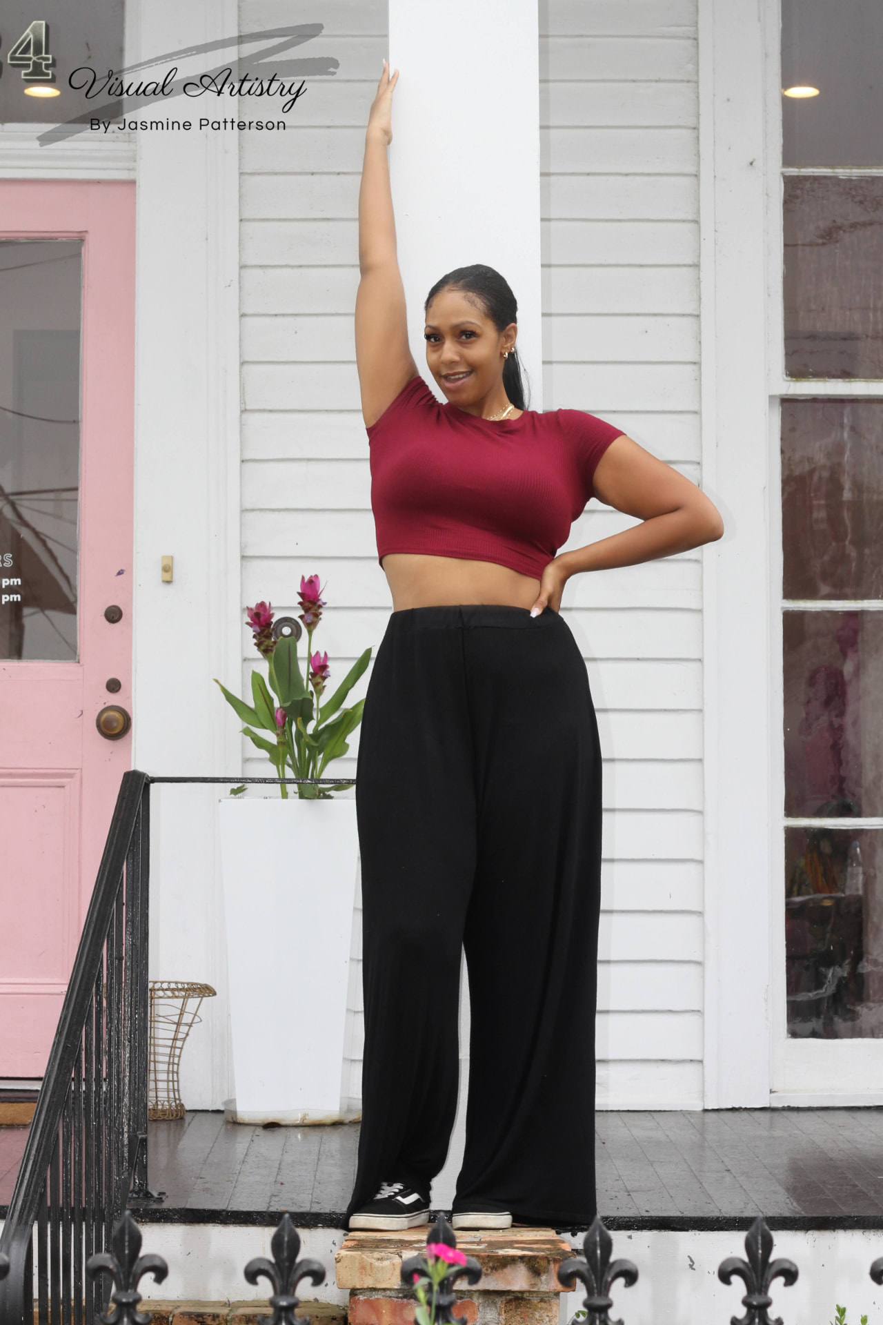 Woman in a burgundy crop top and black trousers posing on a porch beside a pink door
