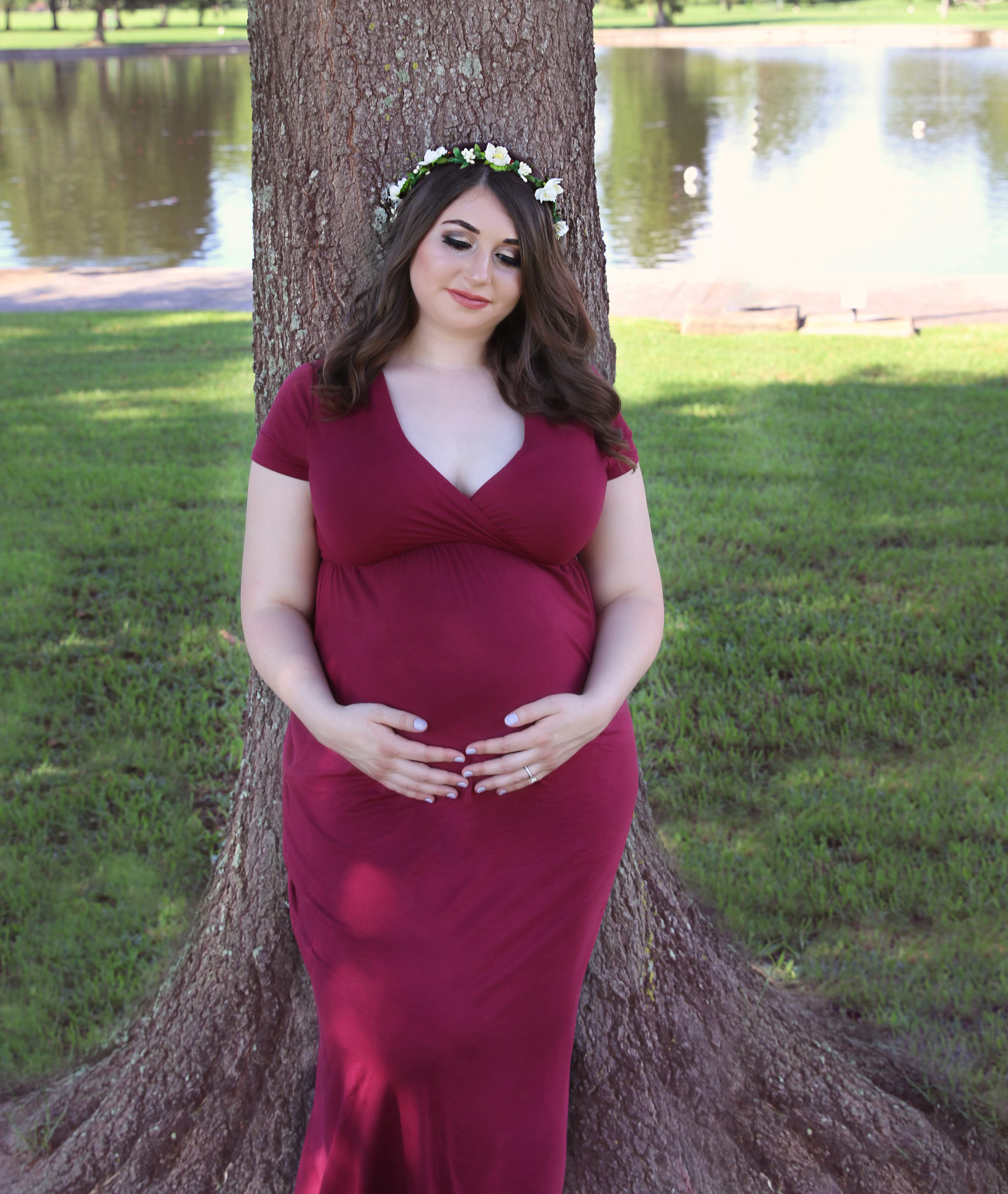 Expectant mother in a burgundy gown and flower crown standing beside a tree
