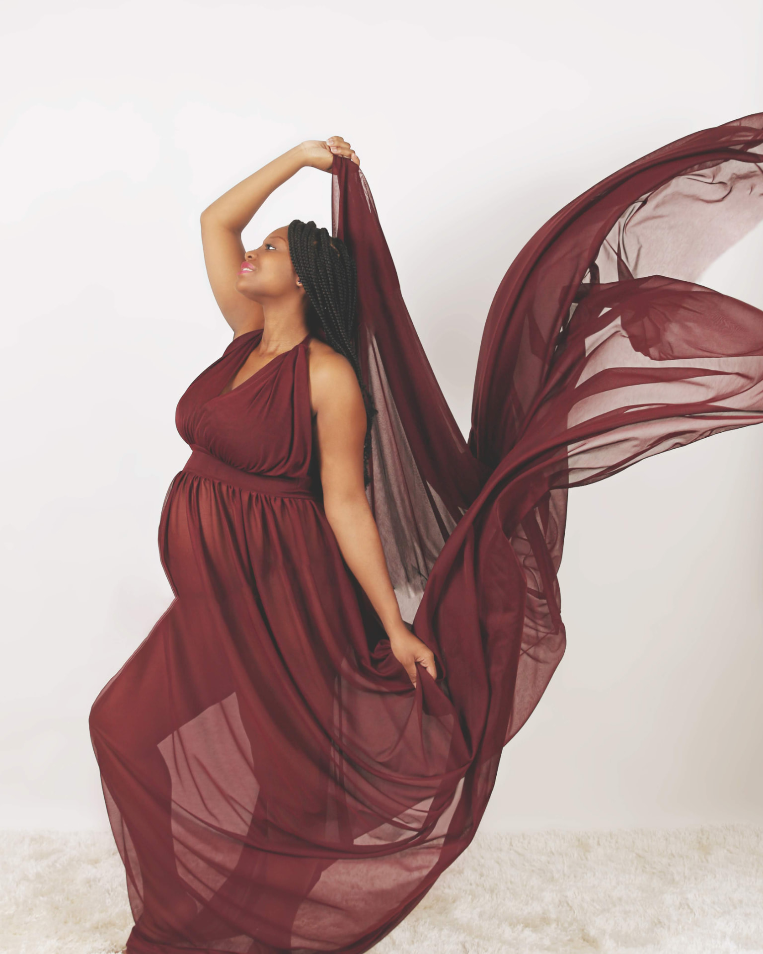 Pregnant woman in a flowing maroon gown lifting sheer fabric overhead in a bright studio