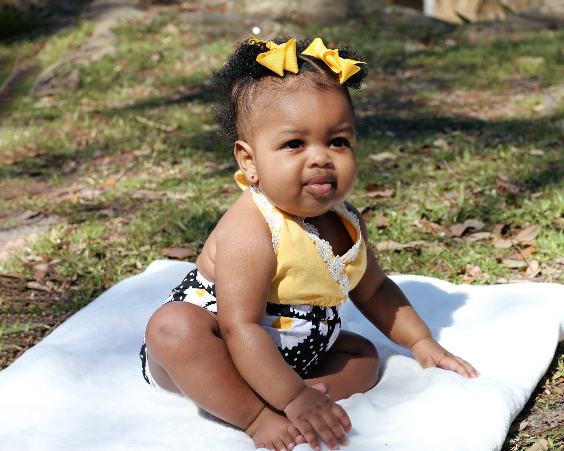 Baby girl with a yellow bow seated on a white blanket in the grass