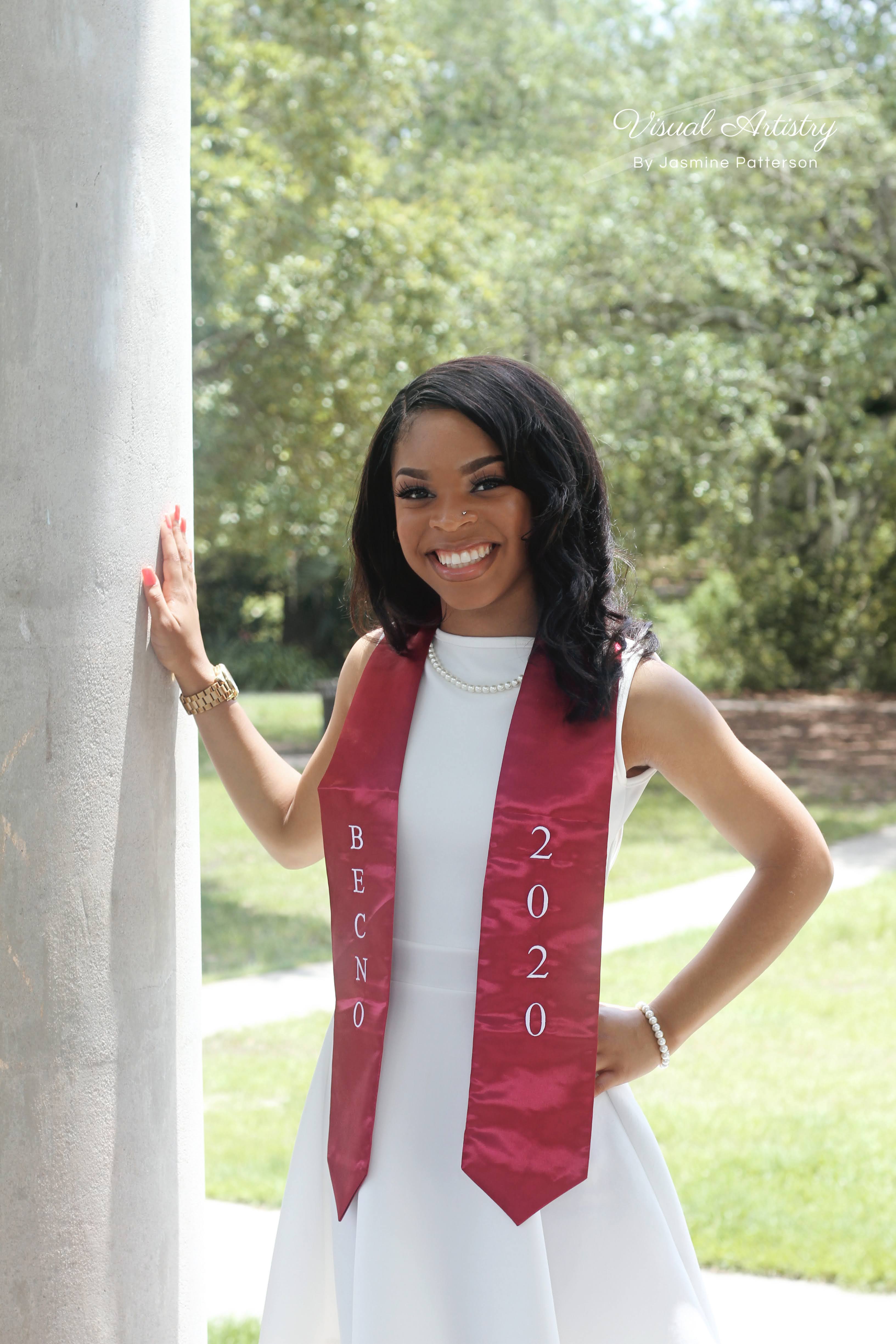 Graduate in a white dress with a red Class of 2020 stole standing by a column