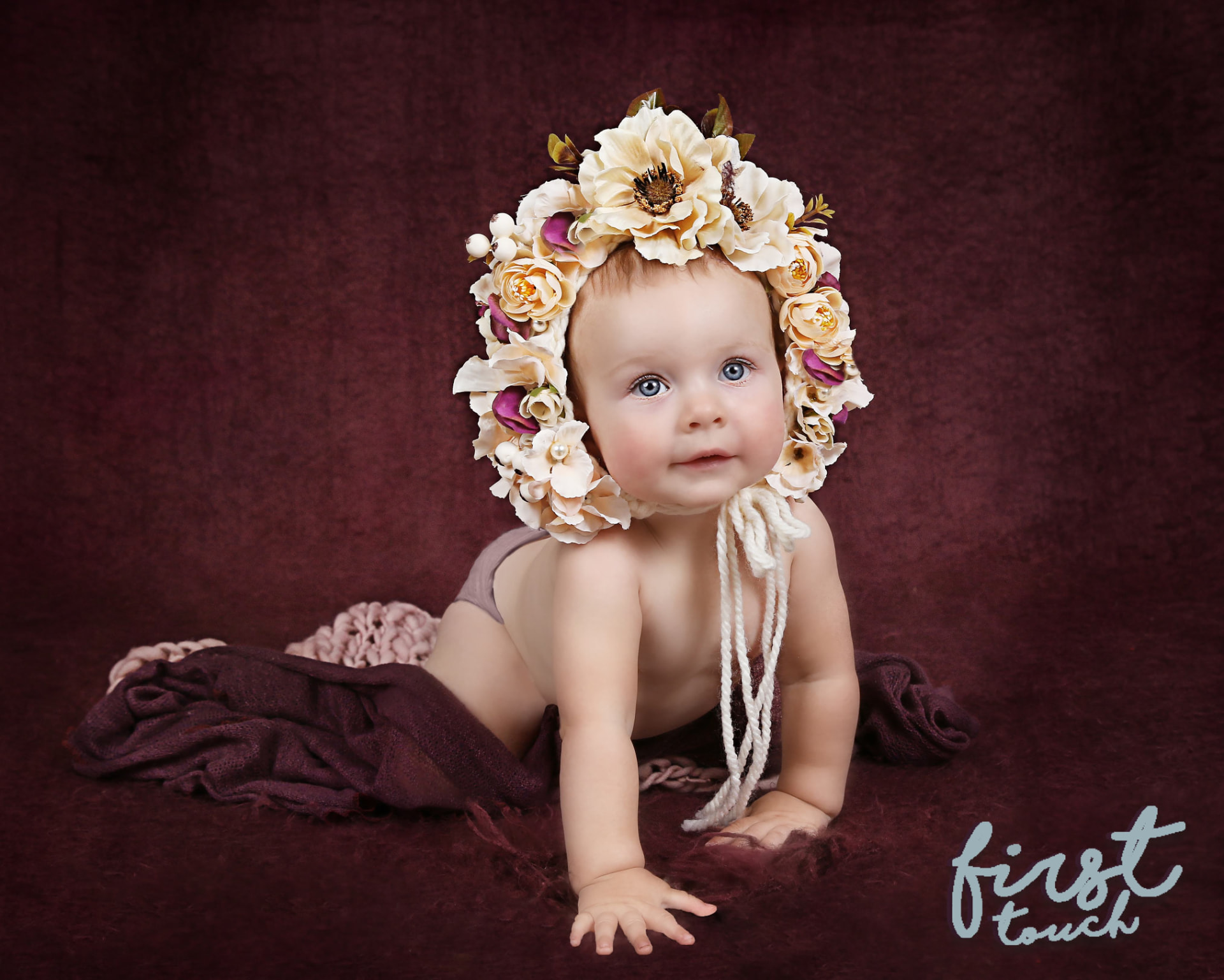 Baby wearing a cream flower bonnet on a deep burgundy backdrop