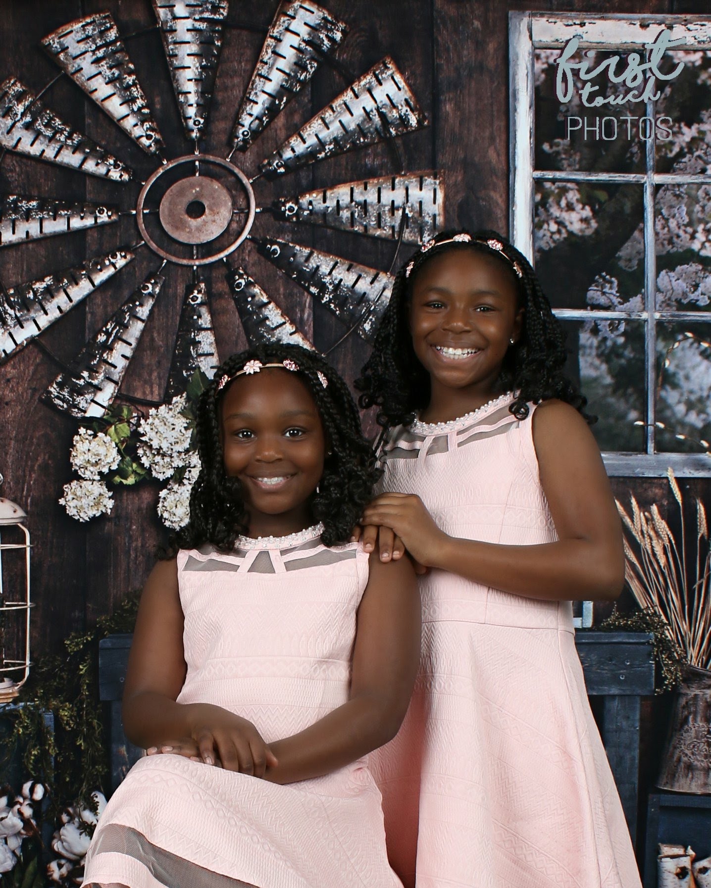 Two sisters in pink dresses with flower crowns before a rustic windmill backdrop