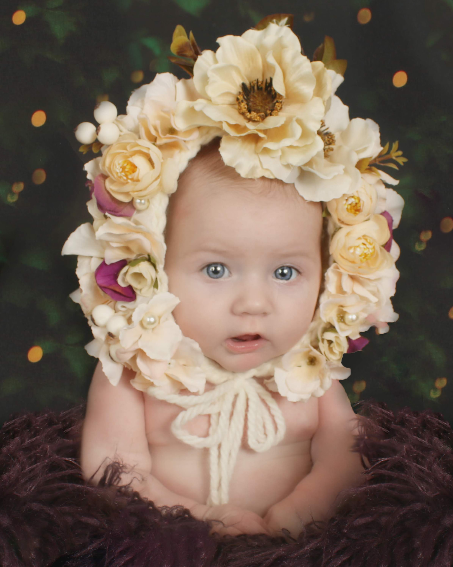 Awake newborn in a cream flower bonnet against a dark feathery backdrop with soft bokeh