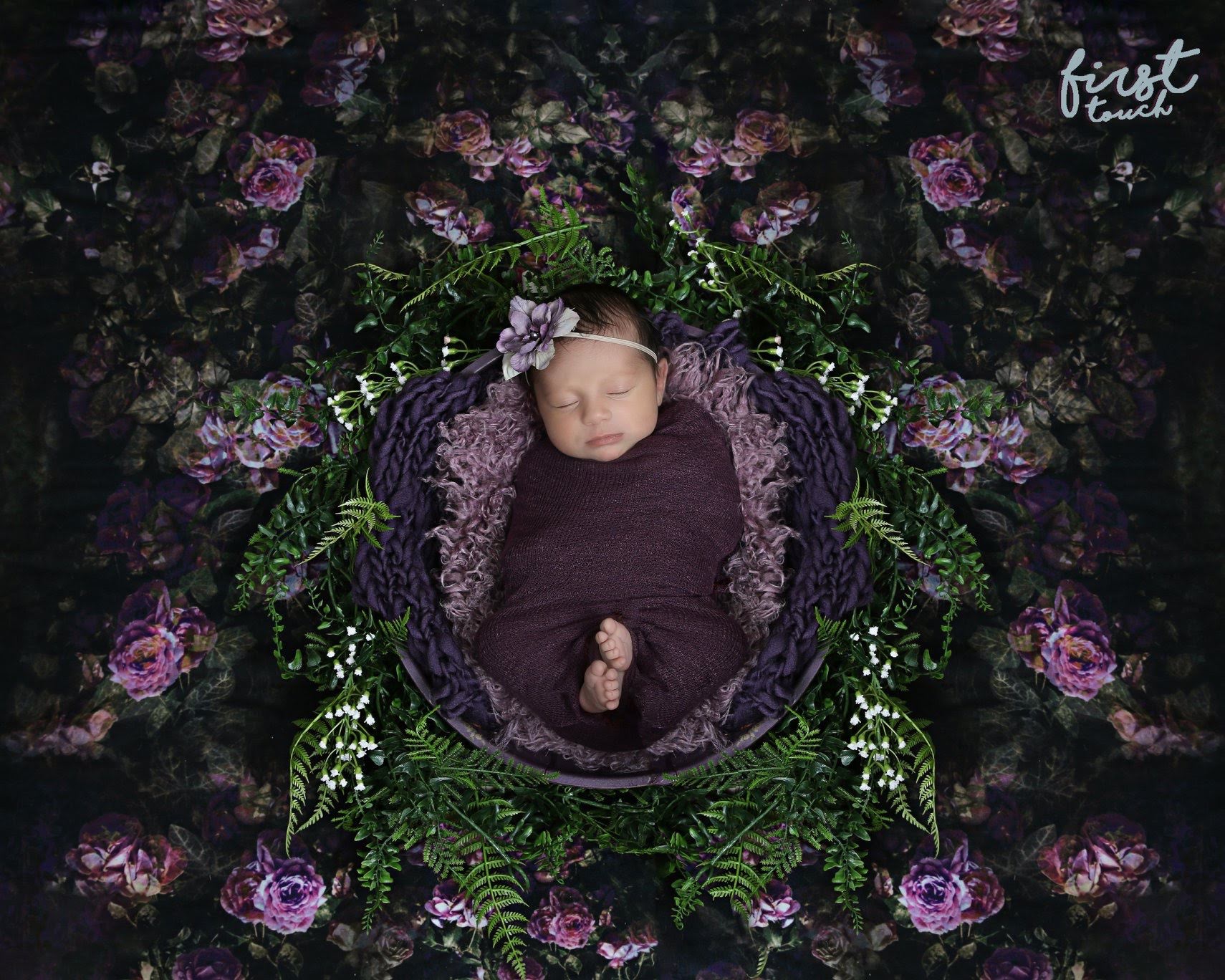 Newborn baby in a purple wrap resting in a greenery-lined bowl surrounded by roses and ferns