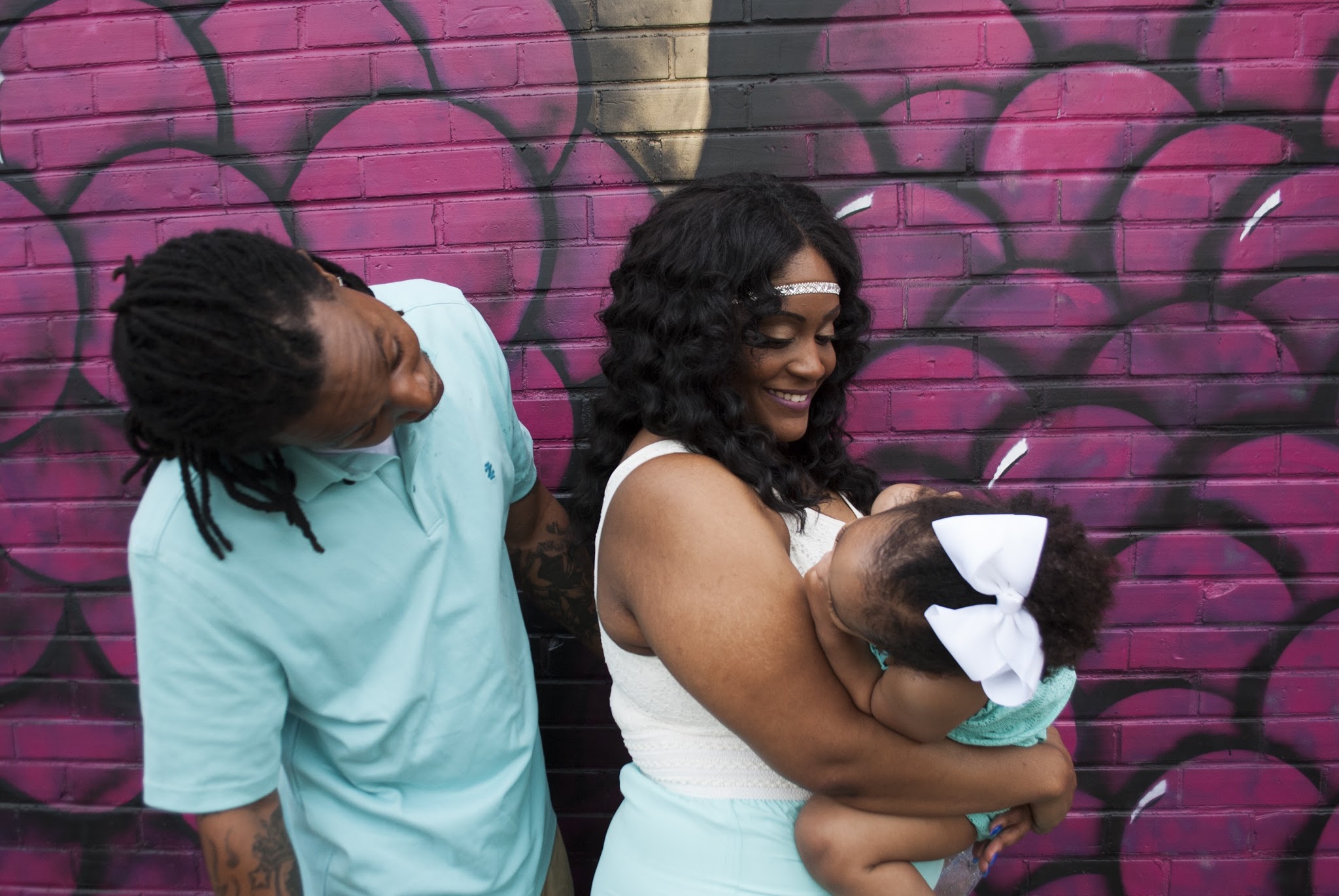 Parents kissing their baby's head in front of a magenta mural wall
