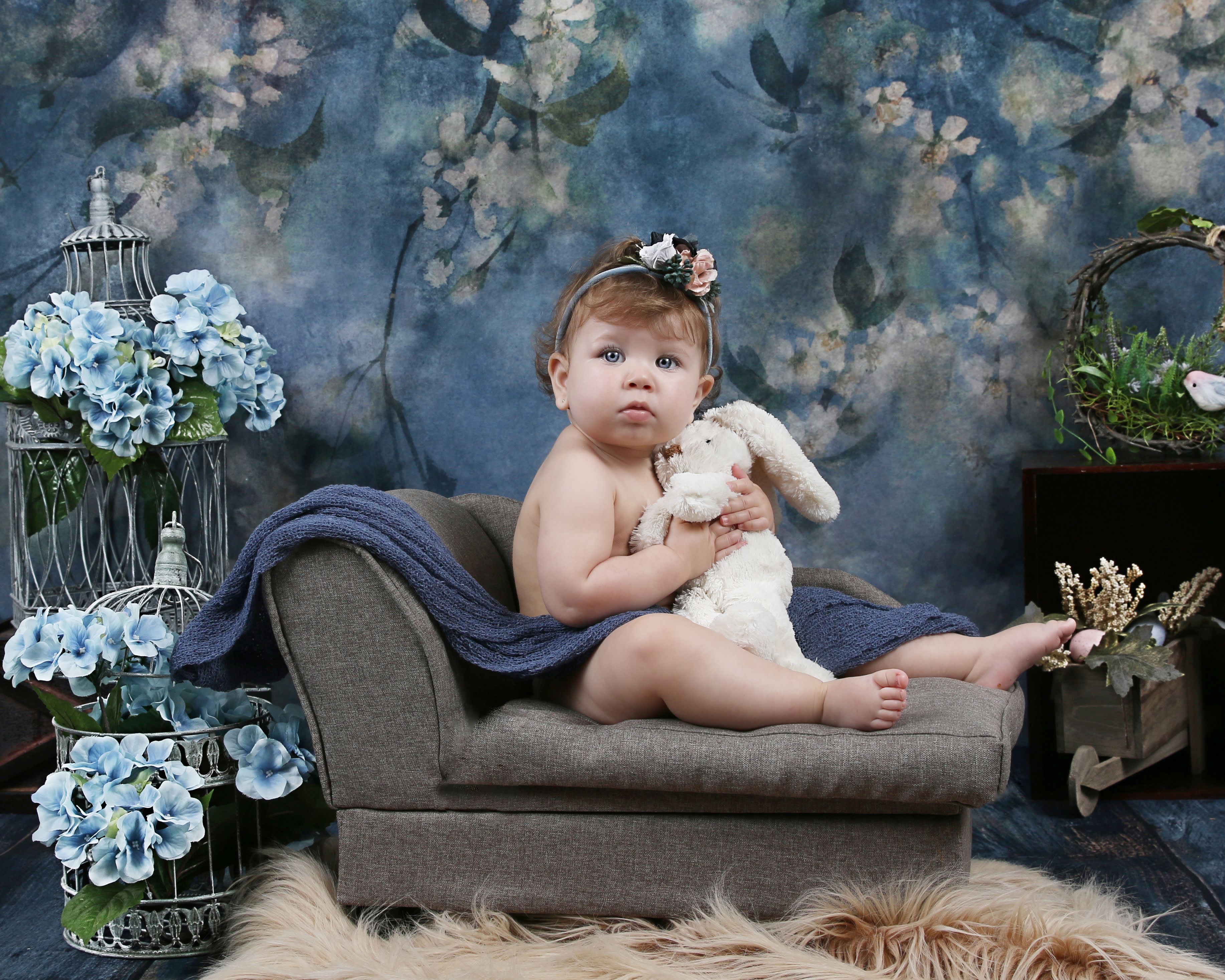 Baby seated on a gray chaise in a blue floral studio set