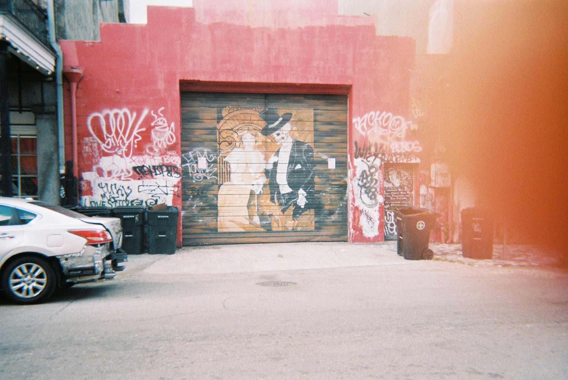 Red-walled alley with a portrait mural painted on a garage door and a warm film light leak