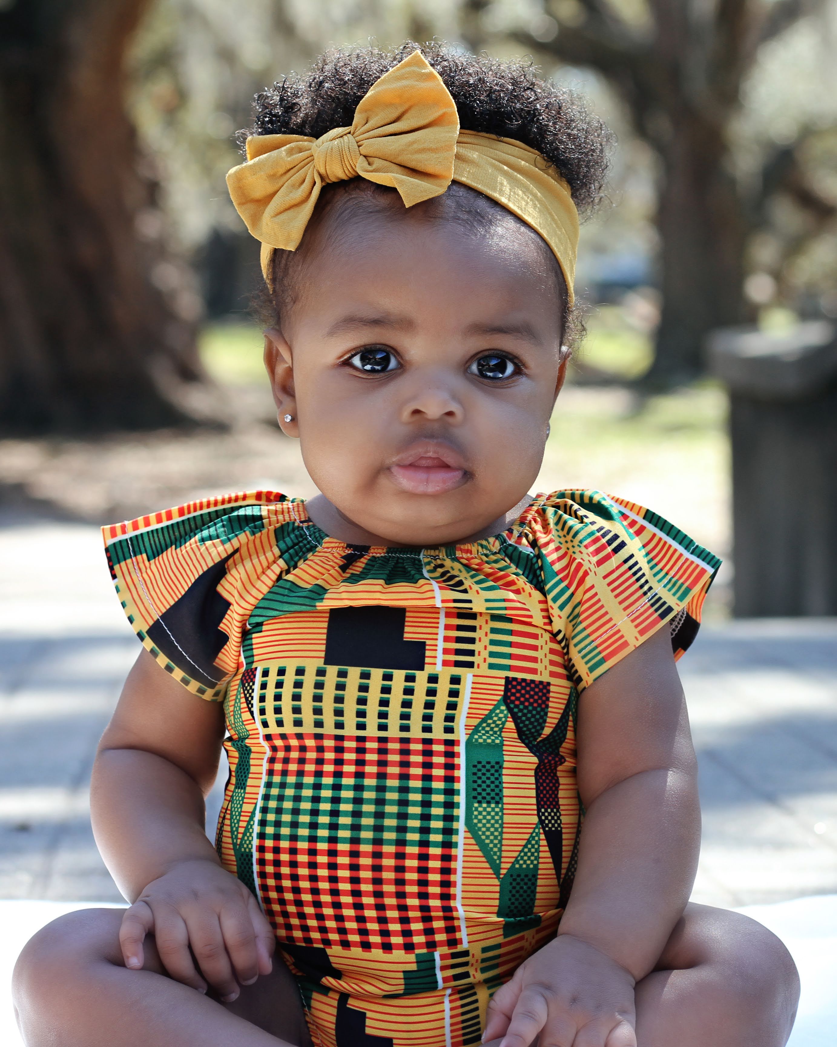 Baby girl in a kente-print outfit with a mustard bow, seated outdoors