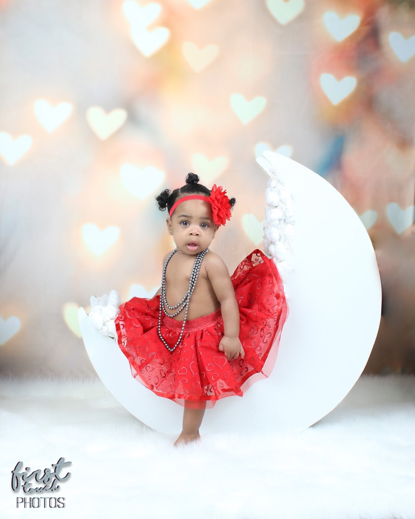 Baby in a red tutu and beads sitting on a white crescent moon against glowing heart bokeh