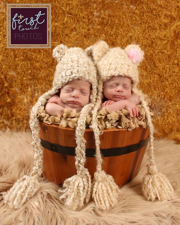 Newborn twins sleeping together in knit bonnets inside a small wooden barrel
