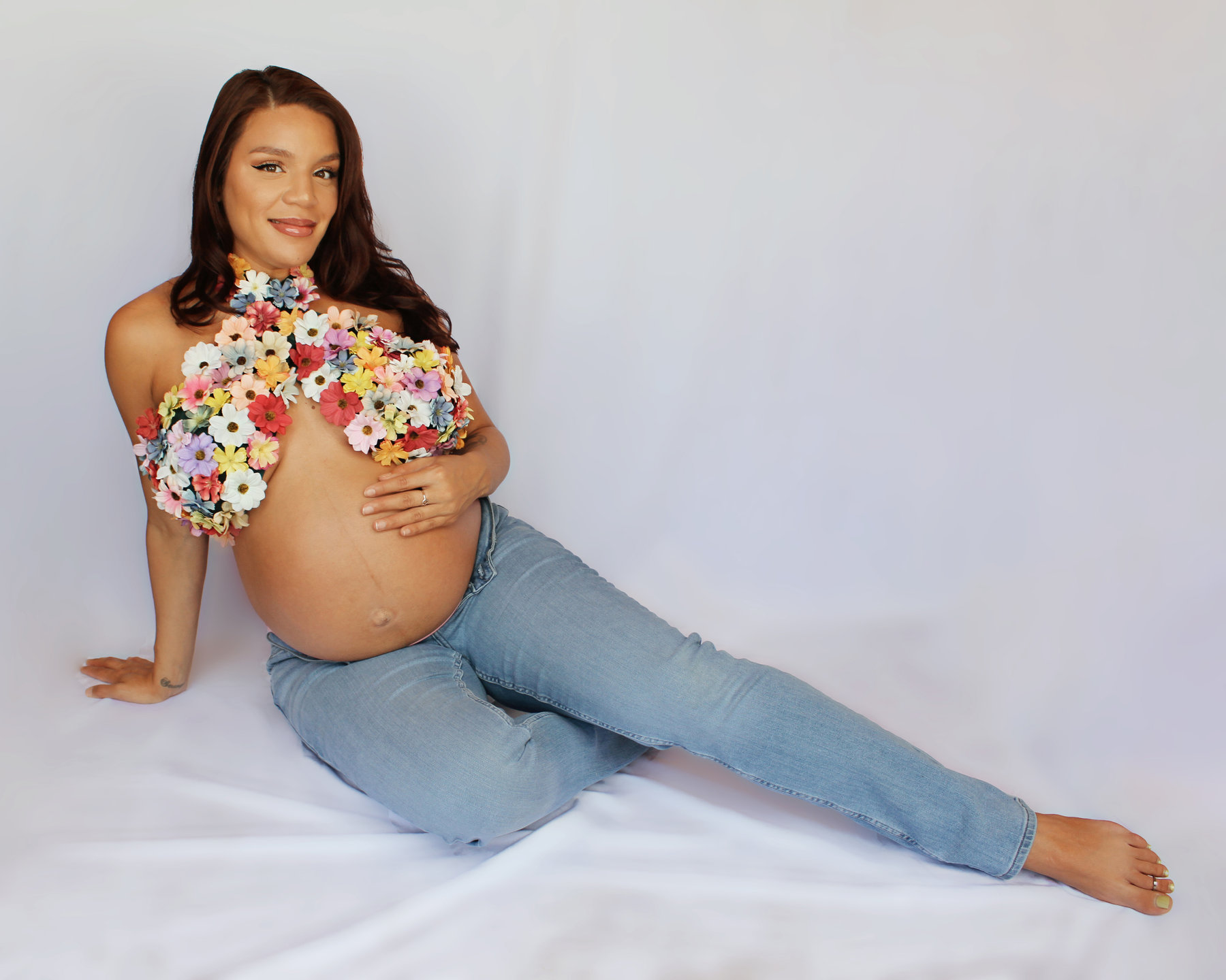Pregnant woman in jeans and a floral top reclining on a white backdrop